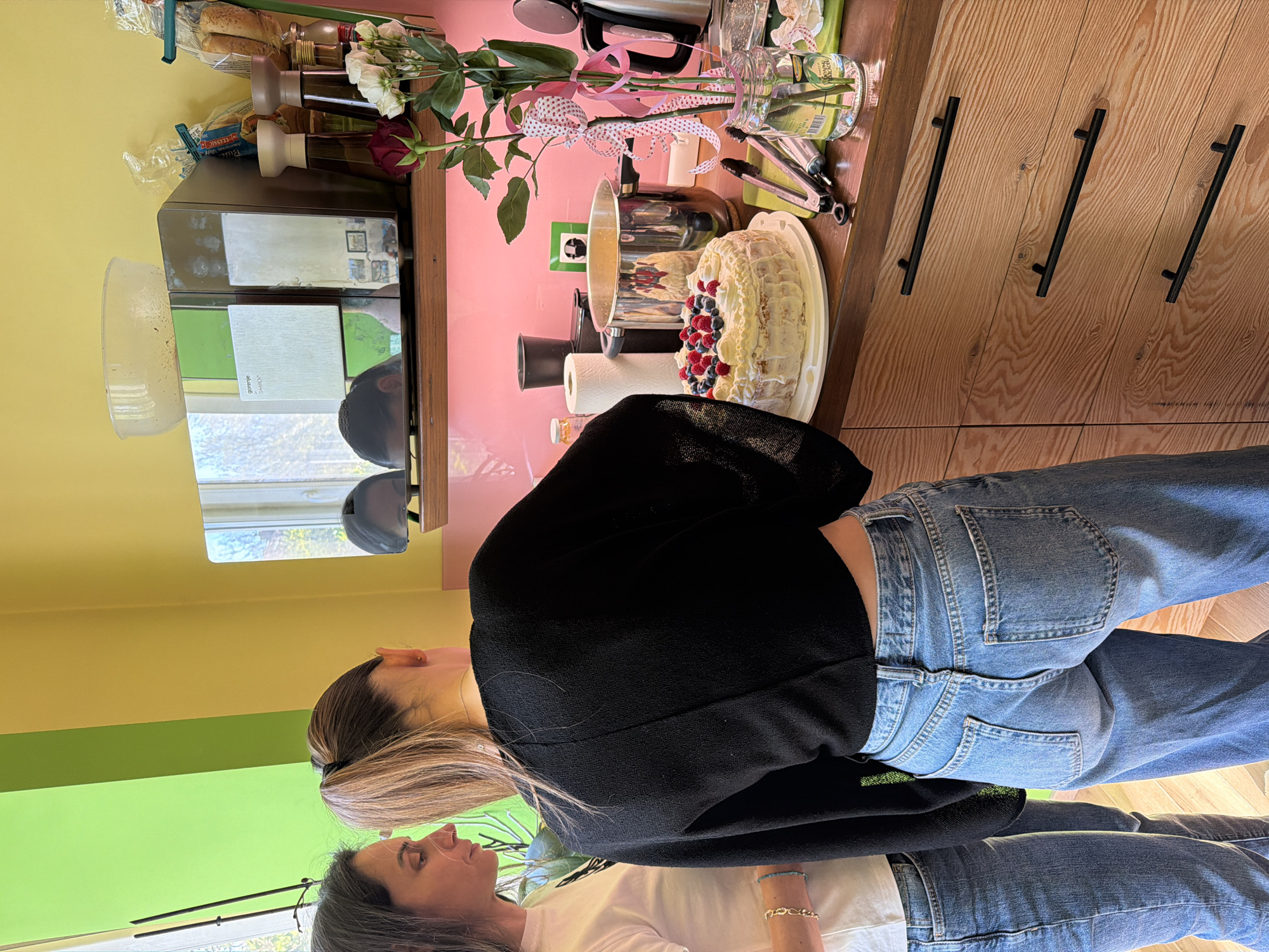 The image shows a brightly lit interior, possibly a kitchen. Two women are present: one with long brown hair in a ponytail, wearing a black jacket and blue jeans, and another woman with grey hair, wearing a white shirt and blue jeans. They appear to be interacting, potentially looking at something on a countertop. On the countertop, there is a cake decorated with white frosting and fresh berries, alongside various kitchen items, including a metal pot, flowers in a glass vase tied with ribbons, and other containers. The background includes a wall painted in yellow and green, and wooden cabinets with black handles. There is a reflection in a window, revealing an outdoor scene, possibly a cityscape. It is not possible to pinpoint the exact location in Sofia, Bulgaria, without additional information.