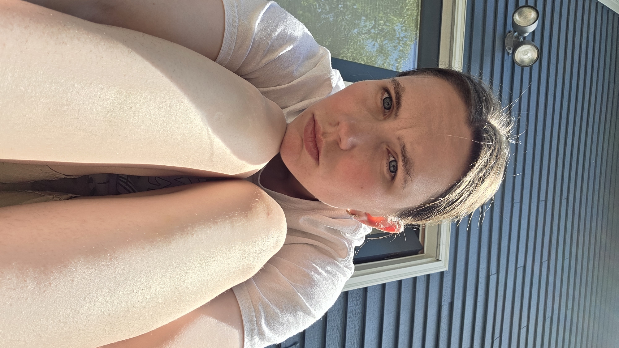 A woman with light skin and brown hair pulled back is sitting with her knees pulled up to her chest, looking directly at the camera. She is wearing a white t-shirt. Her face is illuminated by sunlight, casting shadows on her forehead and nose. Her eyes are blue and her lips are slightly pursed. The background is a dark blue clapboard wall of a house with a light-colored door frame and a window to the left. Two outdoor light fixtures are visible on the wall above her head. The image appears to be taken outdoors in Portland, United States of America.