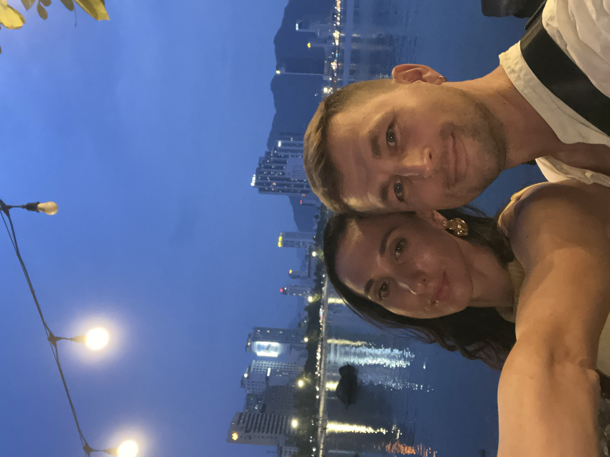 A couple is posing for a selfie at night in the city of Bac Nha Trang Ward, Vietnam. The man has short blond hair and stubble. The woman has dark hair and is wearing gold earrings. Behind them is a body of water reflecting the lights of the city skyline. Streetlights are visible in the foreground.
