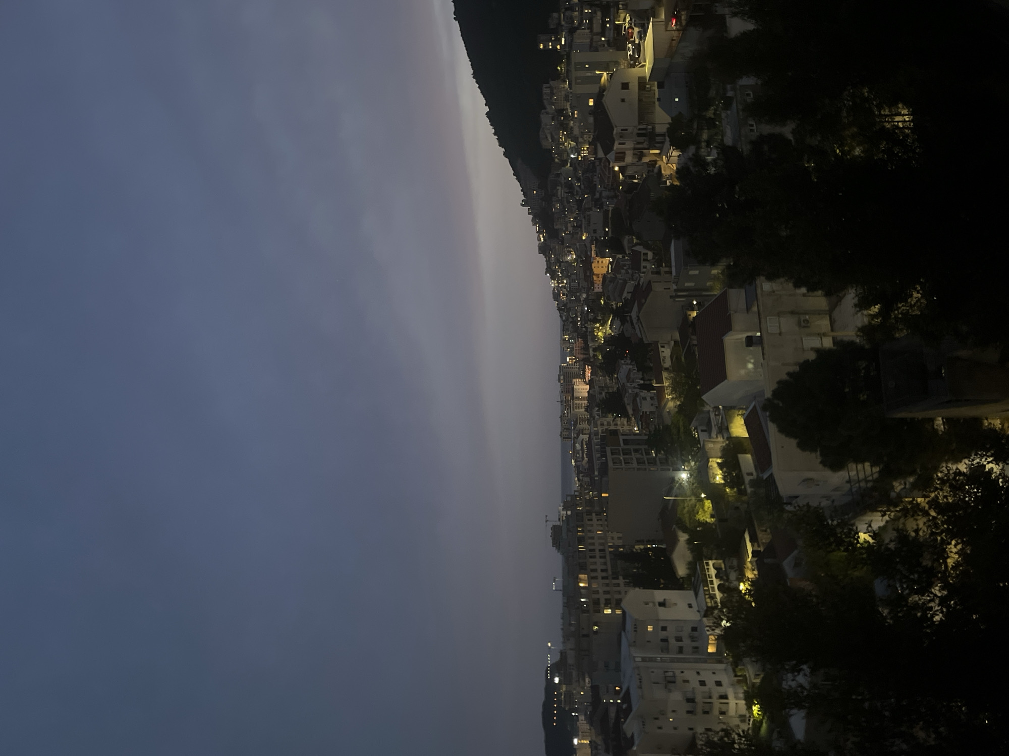 The image depicts a nighttime view of Budva, Montenegro. The sky is a dark, almost black, gradient with a lighter horizon line. A hillside or mountain is visible along the city edge, leading down to a densely packed urban area illuminated by numerous lights from buildings and street lamps. Buildings range in height, creating a tiered effect up the hill. Some trees are visible in the foreground, obscuring parts of the city view.