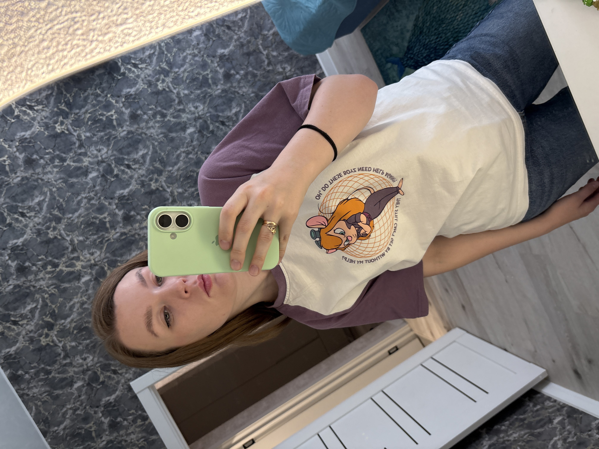 A young woman is depicted taking a mirror selfie in an indoor setting, likely a bathroom or hallway, in Izhevsk, Russia. The image is rotated 90 degrees clockwise.
The woman is visible from the waist up, holding a light green smartphone with her right hand. She is looking directly at the phone's camera, with her lips slightly pursed in a selfie pose. She has long, straight brown hair.
She is wearing a two-toned, long-sleeved t-shirt with a white main body and purple or maroon sleeves. The t-shirt features a graphic design of a cartoon character, possibly a mouse or cat with orange hair, sitting. Mirrored text on the shirt, when reversed, reads: "OH, DO THESE BOYS NEED HELP WITHOUT MY HELP?". She is also wearing blue denim jeans.
Accessories include a gold-toned ring on her right ring finger and a thin black band, likely a hair tie or bracelet, on her left wrist.
The background features a dark gray or black marbled floor with white veins. A linear light fixture is visible at the top left, emitting a warm glow. A white door or cabinet with horizontal paneling is present in the lower right of the frame. The scene appears well-lit, suggesting an indoor, possibly daytime, environment.