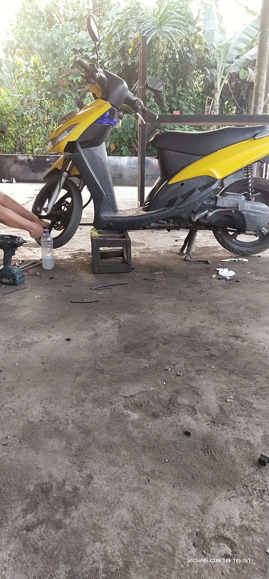 The image depicts an outdoor scene, likely a workshop or garage, with a yellow and black scooter as the primary subject. The scooter is elevated on a dark, square block, suggesting it is being worked on. A person's hands are visible in the lower left corner, holding a water bottle and appearing to be engaged in some activity related to the scooter's front wheel, which is partially obscured by the handlebars and front fairing. A cordless drill is also present on the ground near the hands.
The environment is unpaved, with a dirt or concrete floor that appears dusty and somewhat cluttered with debris, including pieces of white paper or cloth and what might be wire or cable. In the background, lush green foliage suggests a tropical setting, possibly with banana trees and other large-leafed plants. The lighting indicates it is daytime, with sunlight filtering through the leaves, creating bright highlights and shadows. A partial view of a metal structure, possibly a frame or shelving unit, is visible behind the scooter. The timestamp "2026.08.18 16:51" is present in the bottom right corner of the image. The location is identified as Matabu, Indonesia.