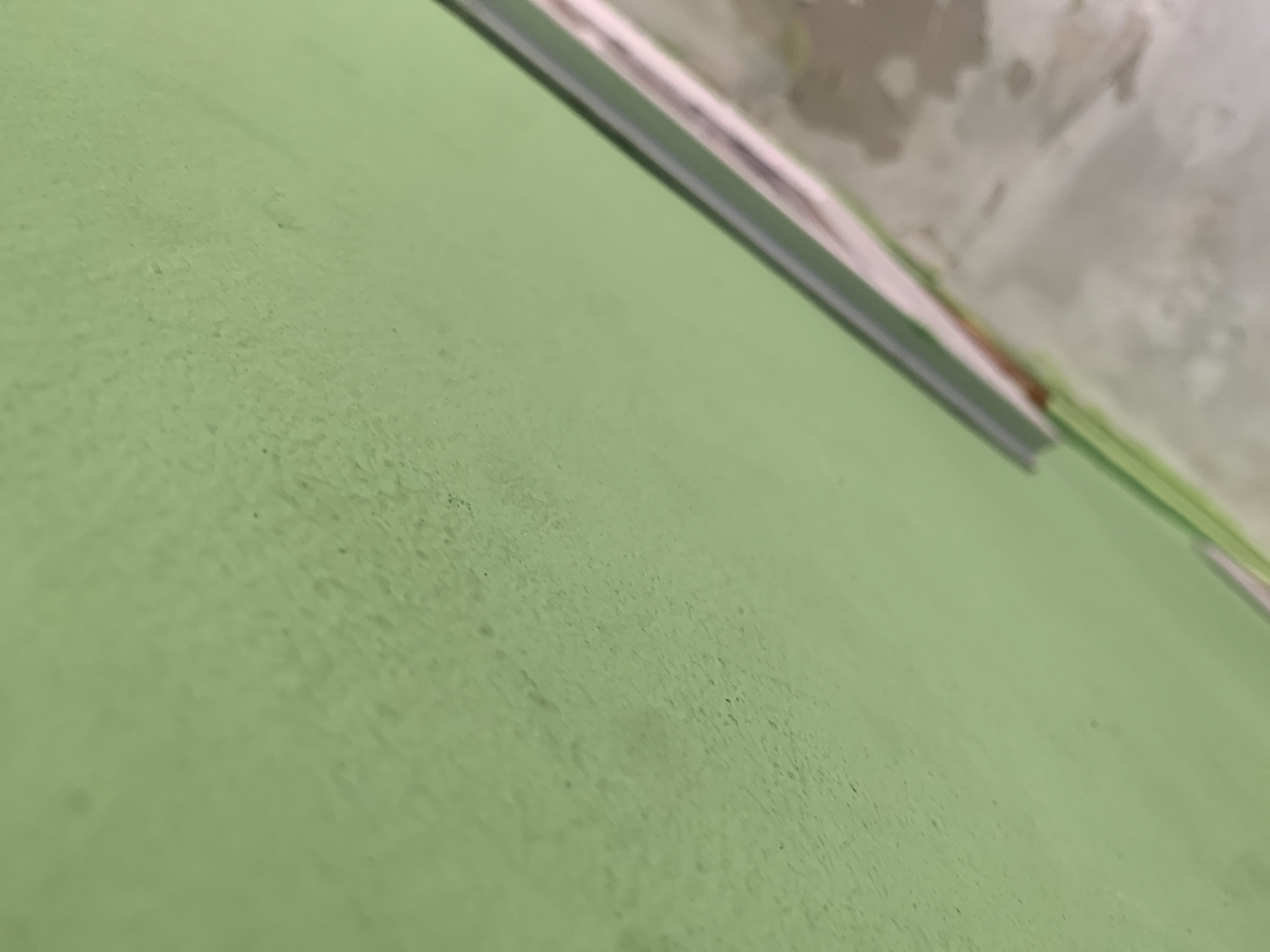 A close-up, angled shot focuses on a pale green surface that appears to be textured. A thin, light-colored object with a metallic edge runs diagonally across the upper right portion of the frame, obscuring a blurred background that suggests a wall with some patchy discoloration or wear. The overall impression is of an indoor, possibly unfinished or work-in-progress, environment. There is no discernible location in Palembang, Indonesia, or any specific details about people, actions, or interactions.