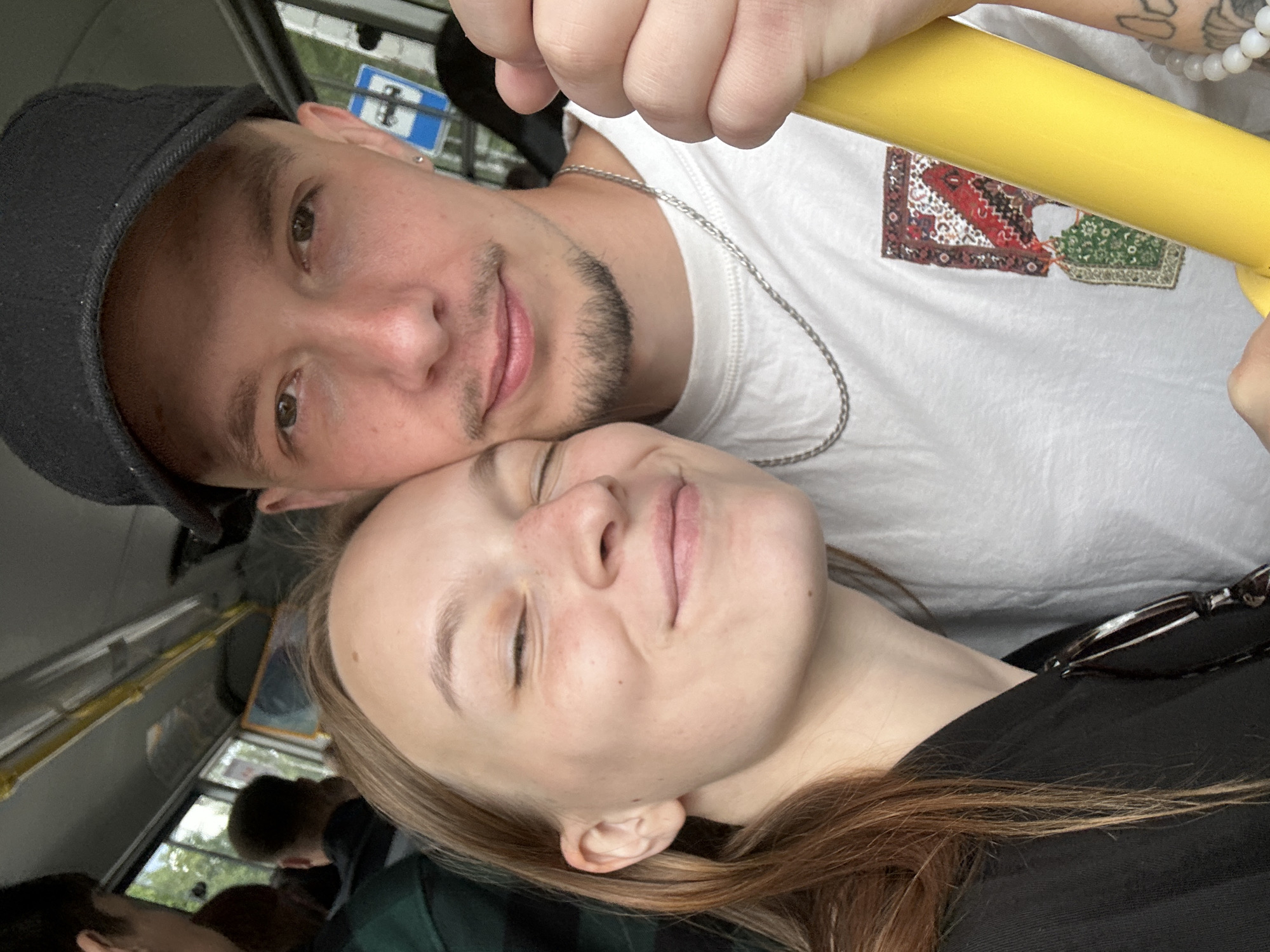 Two people, a man and a woman, are posing for a selfie on what appears to be a public bus. The woman, on the left, has her eyes closed and a smile on her face, with freckles visible on her nose and cheeks. She is wearing a black top. The man, on the right, is wearing a white t-shirt with a patterned patch on the chest and a black baseball cap. He has a mustache and a goatee. They are both looking at the camera. The background shows the interior of the bus, with windows and yellow handrails. A blue traffic sign is visible through the window. The image appears to be taken in Chaikovsky, Russia, based on the presence of a bus which is a common mode of public transportation in the city, and the general aesthetic of the bus interior which is consistent with public transportation in Russia.
