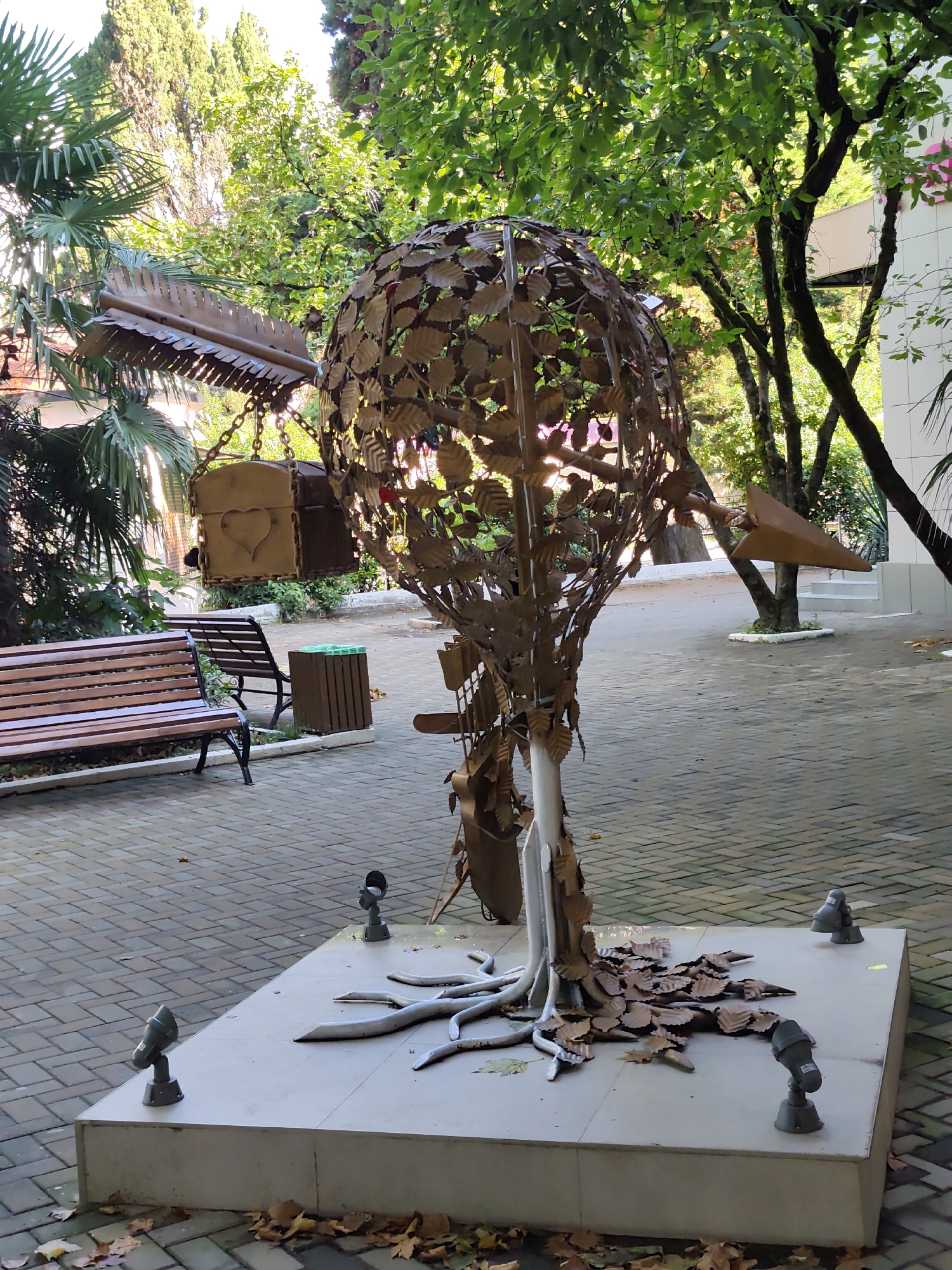 The image shows a public outdoor area with a sculpture of a metallic tree. The sculpture has a spherical canopy made of metallic leaves, and its trunk is supported by metallic root-like structures. At the base of the sculpture, several spotlights are visible. In the background, there is a wooden bench, a trash bin, and lush greenery from palm trees and other deciduous trees, suggesting a warm climate. Part of a building with a white facade is also visible. The ground is paved with interlocking brick tiles. Based on the architectural style, vegetation, and the general ambiance, this scene is consistent with the public spaces found in Sochi, Russia, particularly in resort areas or parks. There are no people or animals visible in the image.