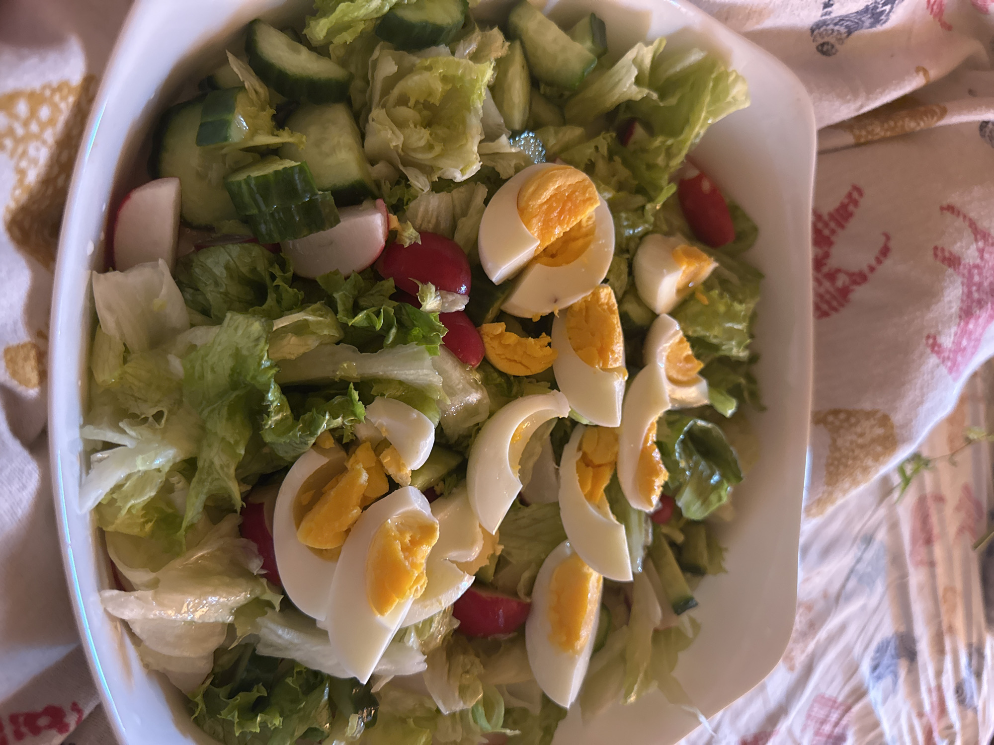 A top-down shot showcases a white, square bowl filled with a mixed salad. The salad consists of lettuce, sliced cucumbers, hard-boiled eggs, and radishes. The bowl sits on a patterned cloth, possibly a tablecloth or napkin. The photograph might have been taken indoors under natural lighting. The city and country are not evident in the image.