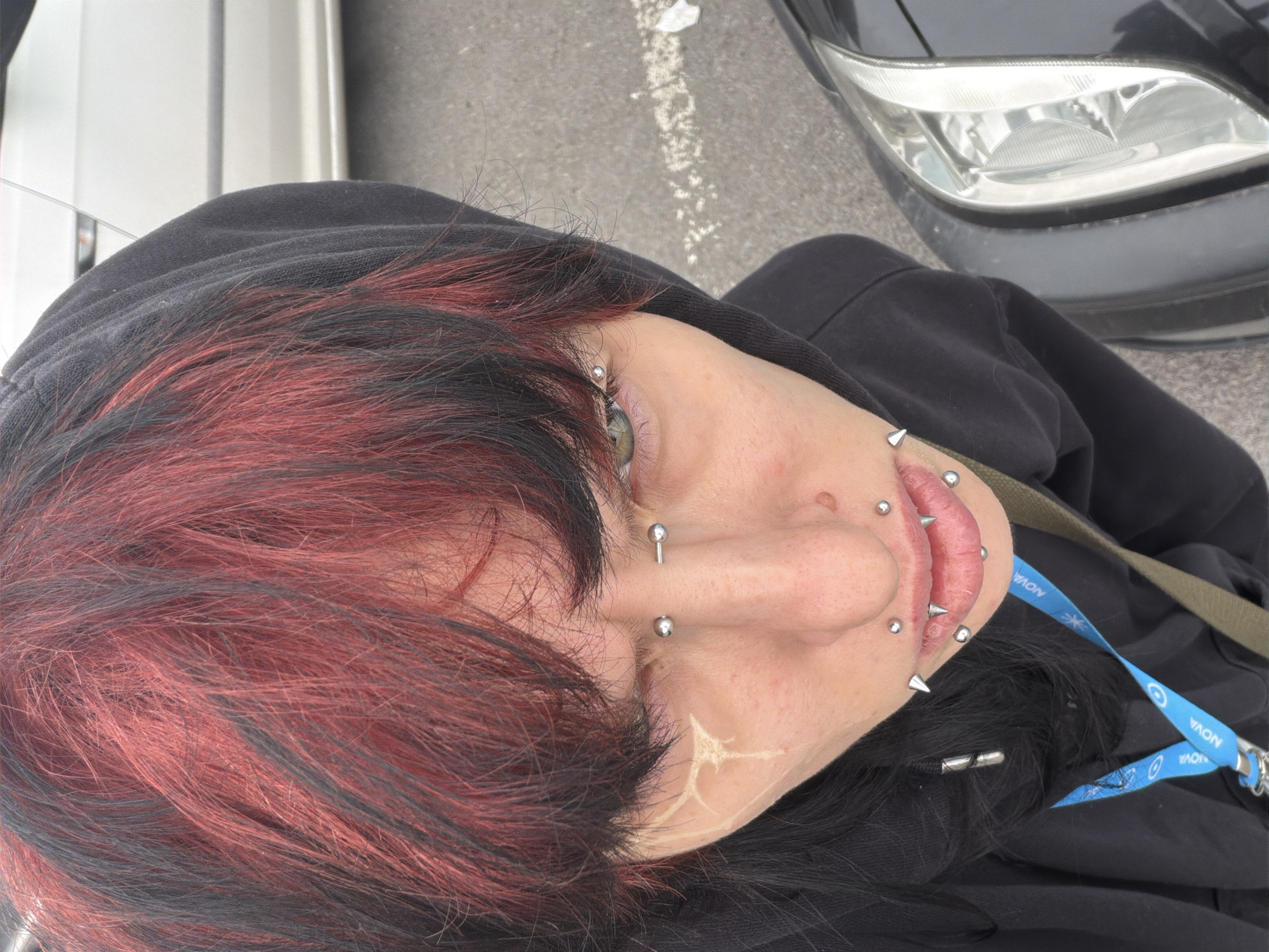 A close-up, angled view shows a person wearing a black hooded sweatshirt. The individual has red and black hair. Multiple facial piercings are visible around the lips, nose, and eyebrow. A lanyard with blue and white text is partially visible near the person's shoulder. A car headlight is present in the upper right corner, and a section of a vehicle is in the upper left. The location is unknown, there are no indications of the city of Sofia, Bulgaria.