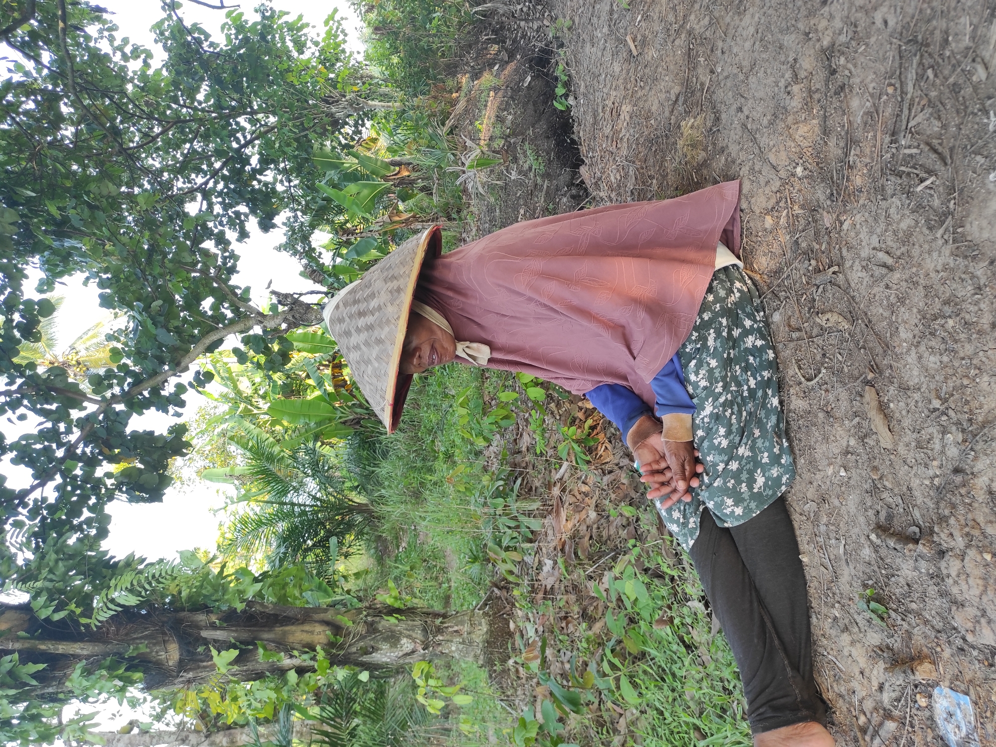 An elderly person wearing a conical Asian hat and a maroon shawl is sitting on the ground next to a dirt embankment in an outdoor environment that appears to be a rural area or overgrown field. The person is wearing a floral-patterned shirt and dark pants. Their hands are clasped in front of them. The background includes green vegetation, trees, and what appears to be a cultivated area. The location is likely somewhere in a rural setting near Lhokseumawe, Indonesia, but it is hard to give an exact location based solely on the provided image.