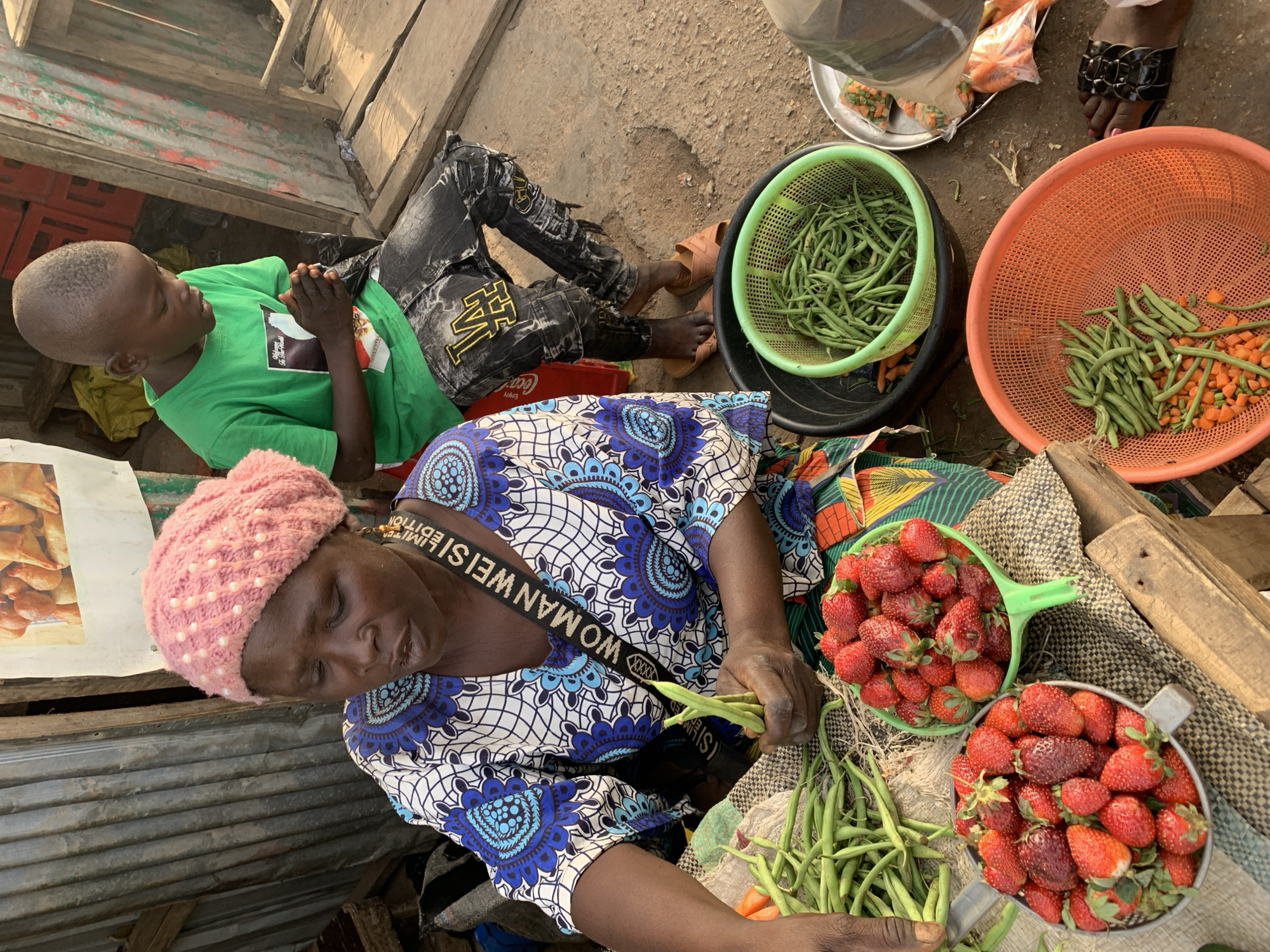 The image shows a street market scene in Jos, Nigeria. An adult woman wearing a blue and white patterned dress and a pink knitted hat is seated on the ground, preparing green beans. She is surrounded by baskets and containers filled with produce including green beans, carrots, and strawberries. A young boy wearing a green t-shirt and black pants is lying on the ground near her. Other individuals and additional baskets are visible in the periphery, along with basic market infrastructure like tin walls.
