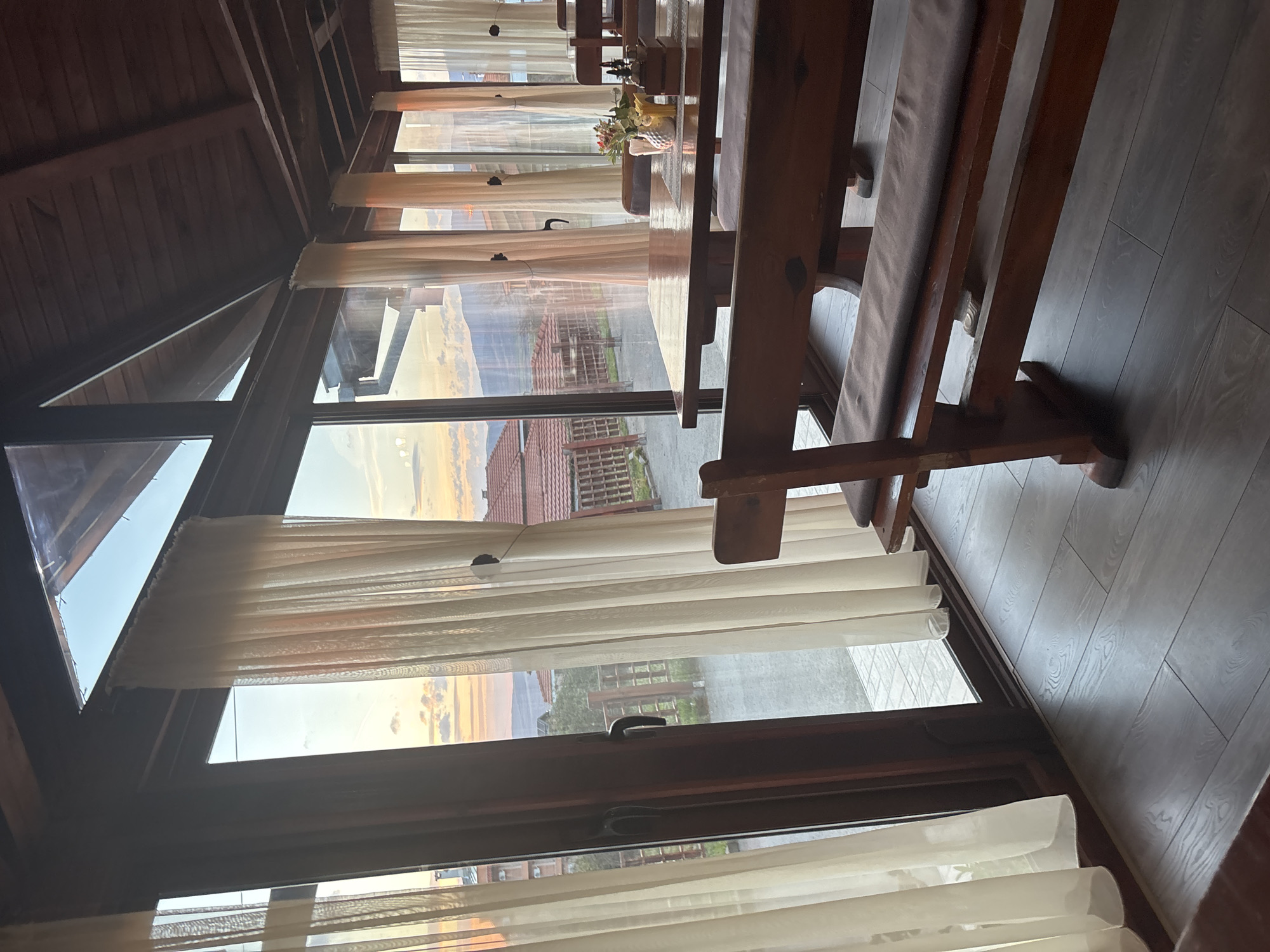 The image shows the interior of a wooden structure, possibly a restaurant or enclosed porch, located in or near Sapareva Banya, Bulgaria. The room features long wooden tables and benches, with light-colored curtains hanging along a series of windows. Through the windows, a landscape is visible with buildings and greenery, with a sky displaying the colors of sunset or sunrise. The floor is made of dark wood planks.