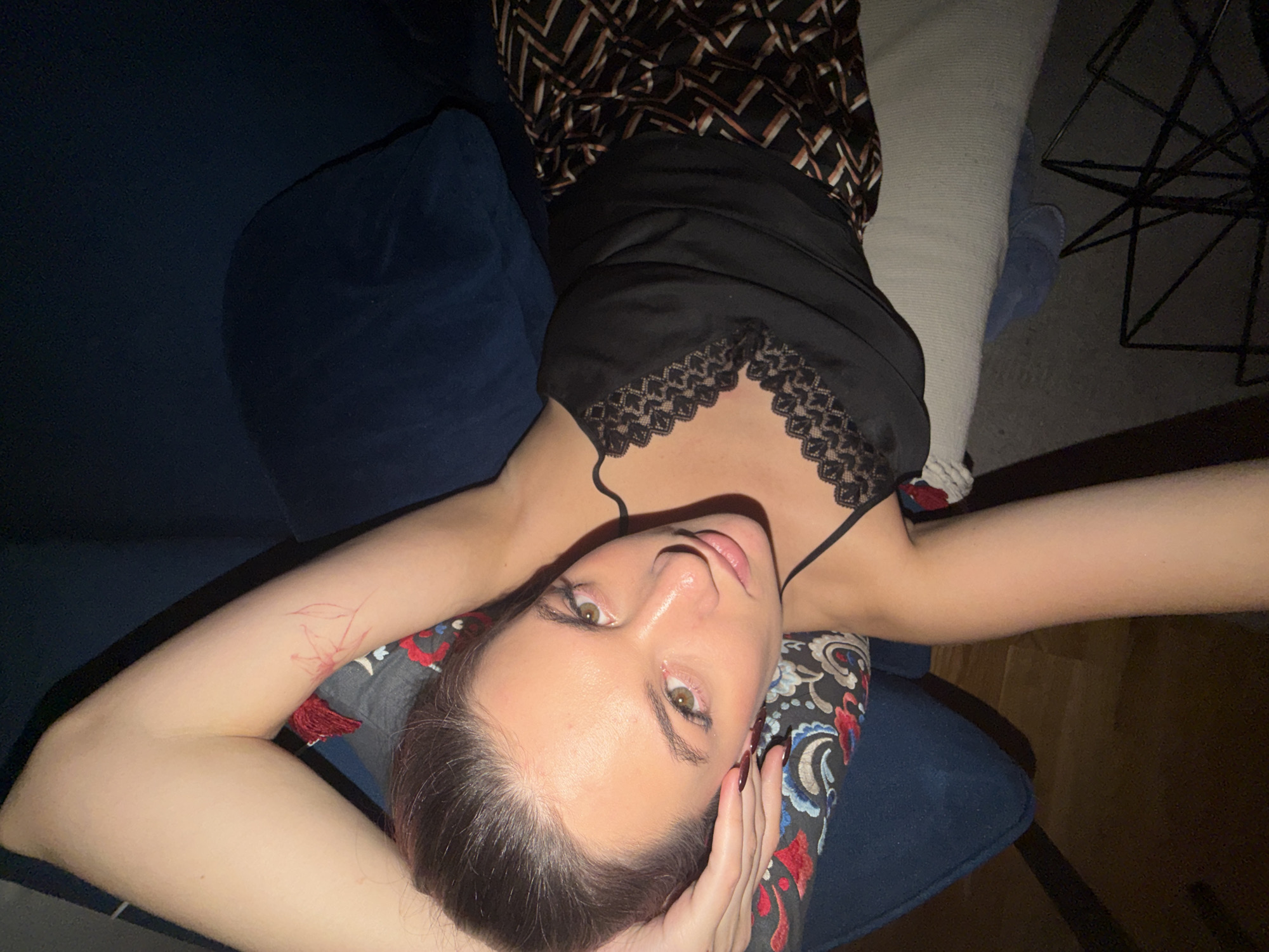 The image displays a young adult female lying supine on an upholstered surface in an interior residential environment located in Belgrade, Serbia. Her head is angled towards the viewer, making direct eye contact with hazel eyes. She has light skin and dark hair, with subtle eye makeup. The subject wears a black lace-trimmed camisole and patterned trousers featuring brown, black, and light-colored geometric designs. A red, hand-drawn floral or leaf pattern is visible on the upper part of her right arm. Her left hand, with dark fingernails, is positioned under her head, resting on a pillow with a multi-colored paisley or floral design in red, blue, and white. Adjacent to her head is a solid dark blue upholstered surface. The floor in the lower right quadrant is dark wood, presenting a black, angular metal object consistent with a lamp base, and a partially obscured light blue slipper beneath the furniture.