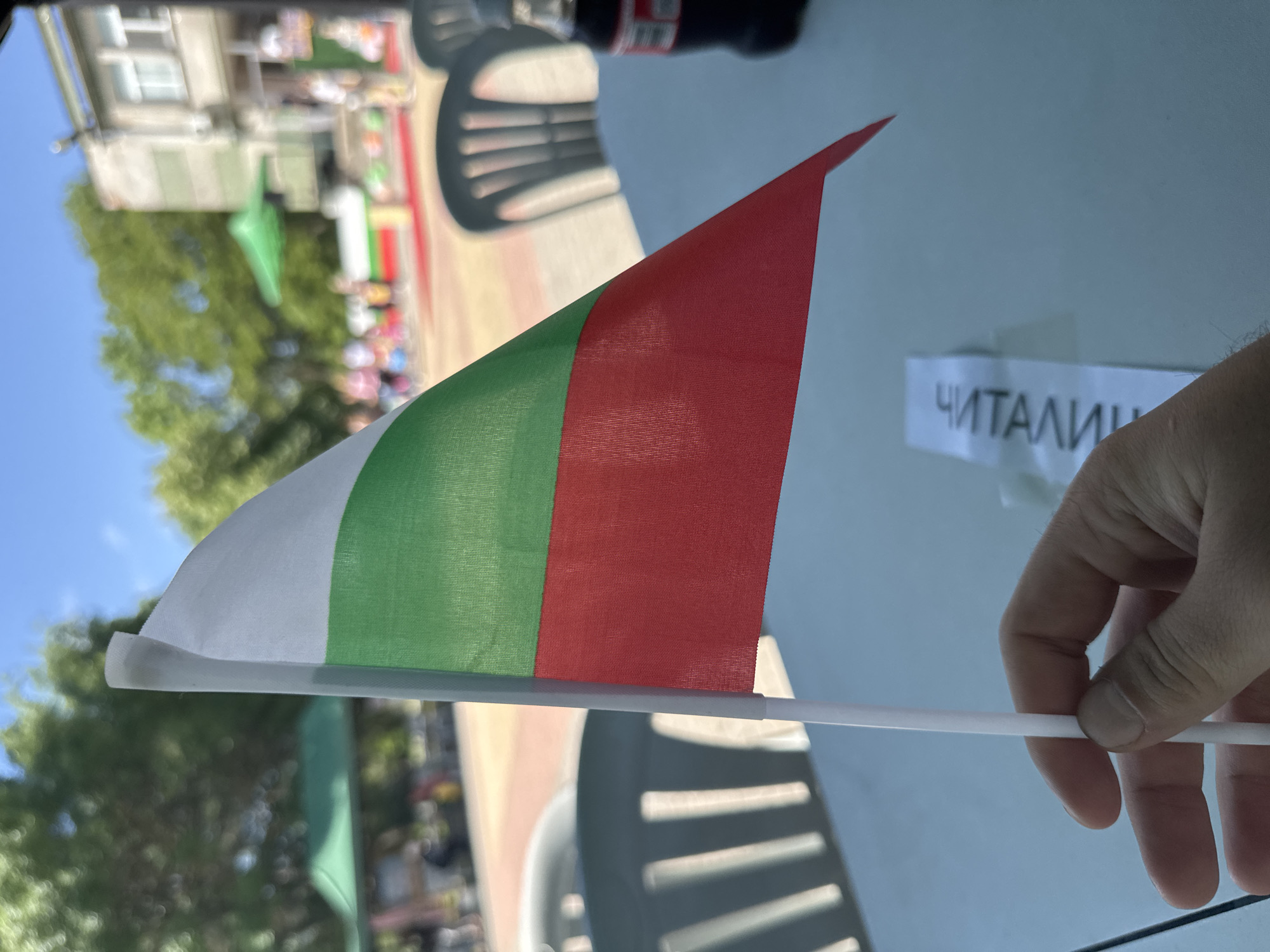 A hand holds a small, pointed Bulgarian flag with white, green, and red stripes. A small white card is visible with the word "читалище" written on it. The background includes blurred images of a building, trees, chairs, tables, and other people, suggesting an outdoor public event or gathering in a city park, which is most likely the city of Novoselets, Bulgaria.