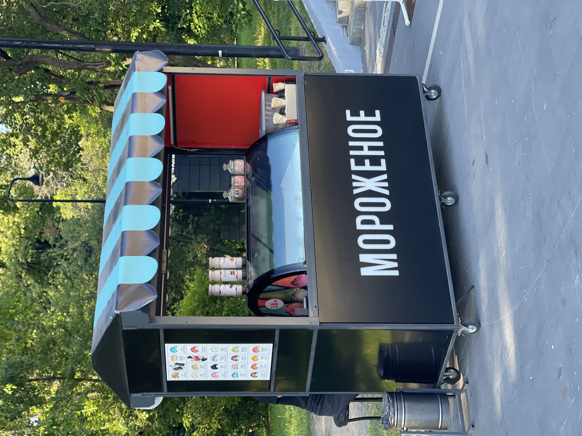 An outdoor ice cream kiosk is shown with a black exterior and a blue and black striped awning. The kiosk has wheels, indicating it is mobile. On the front of the kiosk, the word "МОРОЖЕНОЕ" is written in large white letters. Inside the display case, several tubs of ice cream are visible. To the left of the display case, there is a menu board showing various ice cream flavors. In front of the kiosk, there are two trash cans. A person in dark clothing is sitting on a stool to the left of the kiosk. The kiosk is situated on a paved area, with trees and a lamppost visible in the background. Stairs and a railing are present to the right of the kiosk.