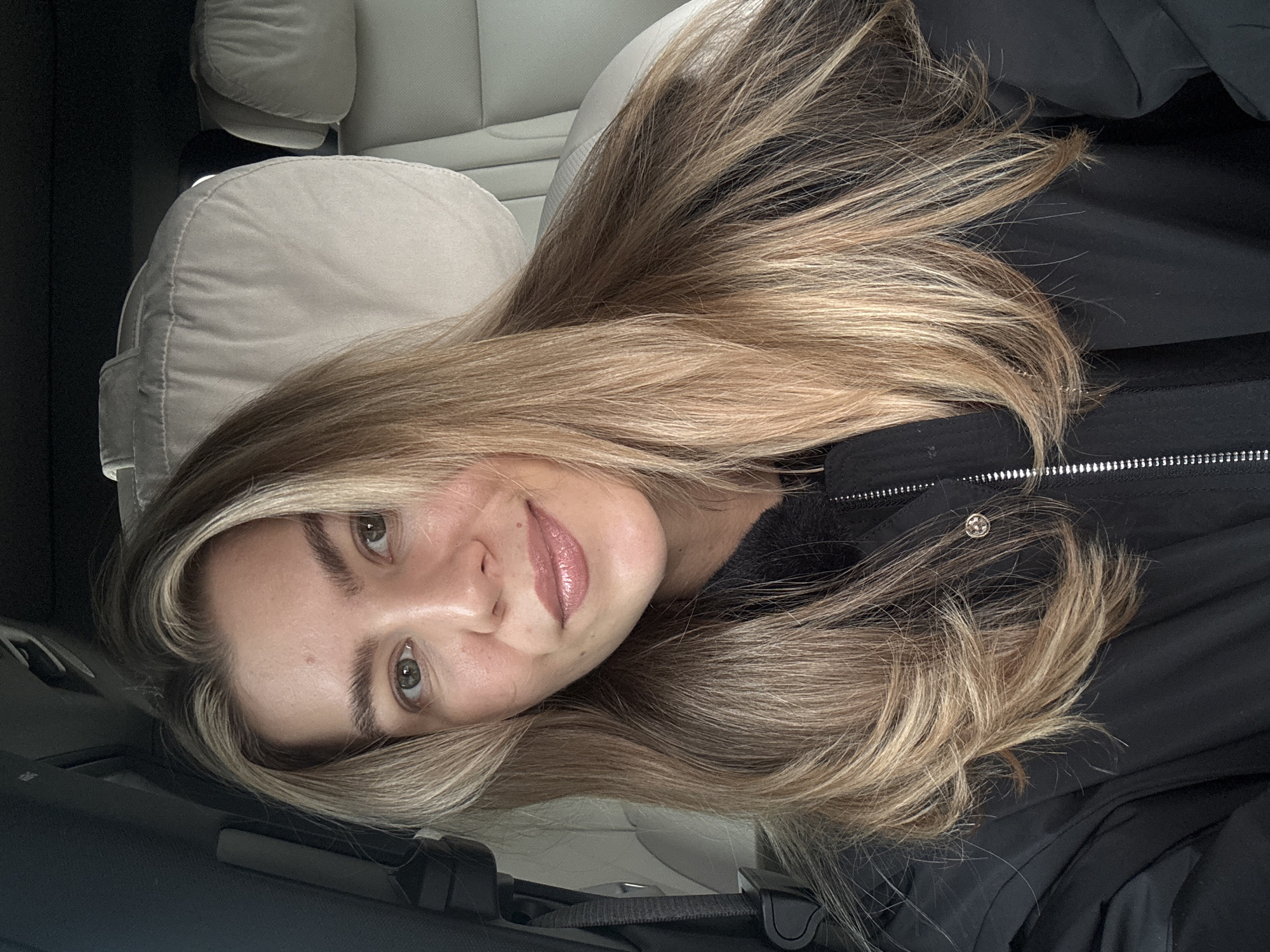 A woman with long, blonde hair is reclined in a vehicle. Her head rests against a light-colored headrest. She is wearing a black jacket. The location is unknown.