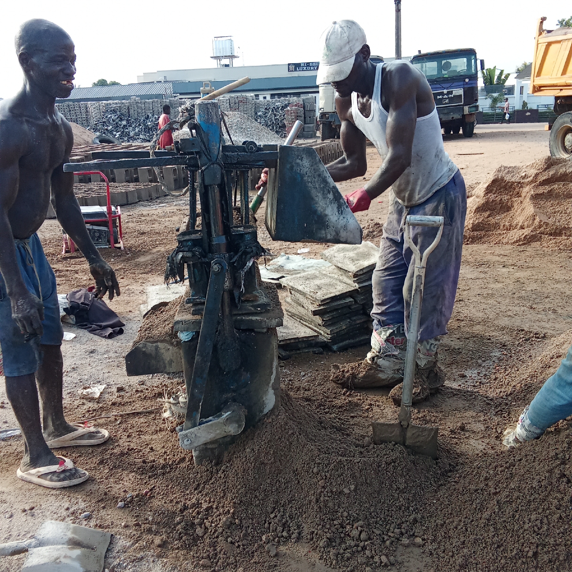 This image captures an outdoor block-making operation, likely at a construction material production site. Two dark-skinned men are prominently featured in the foreground, engaged in manual labor.
The man on the left is bare-chested, wearing blue shorts and white flip-flops. His body and face are covered in dirt, and he is looking towards the viewer with a wide smile, showing his teeth. He appears to be observing or waiting.
The man on the right, wearing a white tank top, blue trousers, a white baseball cap, and red gloves, is actively operating a manual block-making machine. He is captured in the motion of pouring a mixture, likely sand and cement, into the machine's hopper using a metal scoop. His trousers are tucked into what appear to be protective coverings around his ankles, possibly made from sacks or cloth. A shovel stands upright in a pile of material near him.
The ground is dry, reddish-brown earth, with several piles of sand or aggregate. Behind the workers, numerous stacks of freshly made or curing concrete blocks are visible. In the mid-ground, a red metallic frame is partially seen, possibly another piece of equipment or a cart.
In the background, under a bright, clear sky, there are industrial-looking buildings. One building features a sign that reads "RI-SEU LUXURY." Several large vehicles are also present, including a blue truck with the "MAN" logo visible on its grille, and a yellow dump truck. The scene depicts a bustling work environment under daylight conditions, suggesting a focus on the production of building materials.