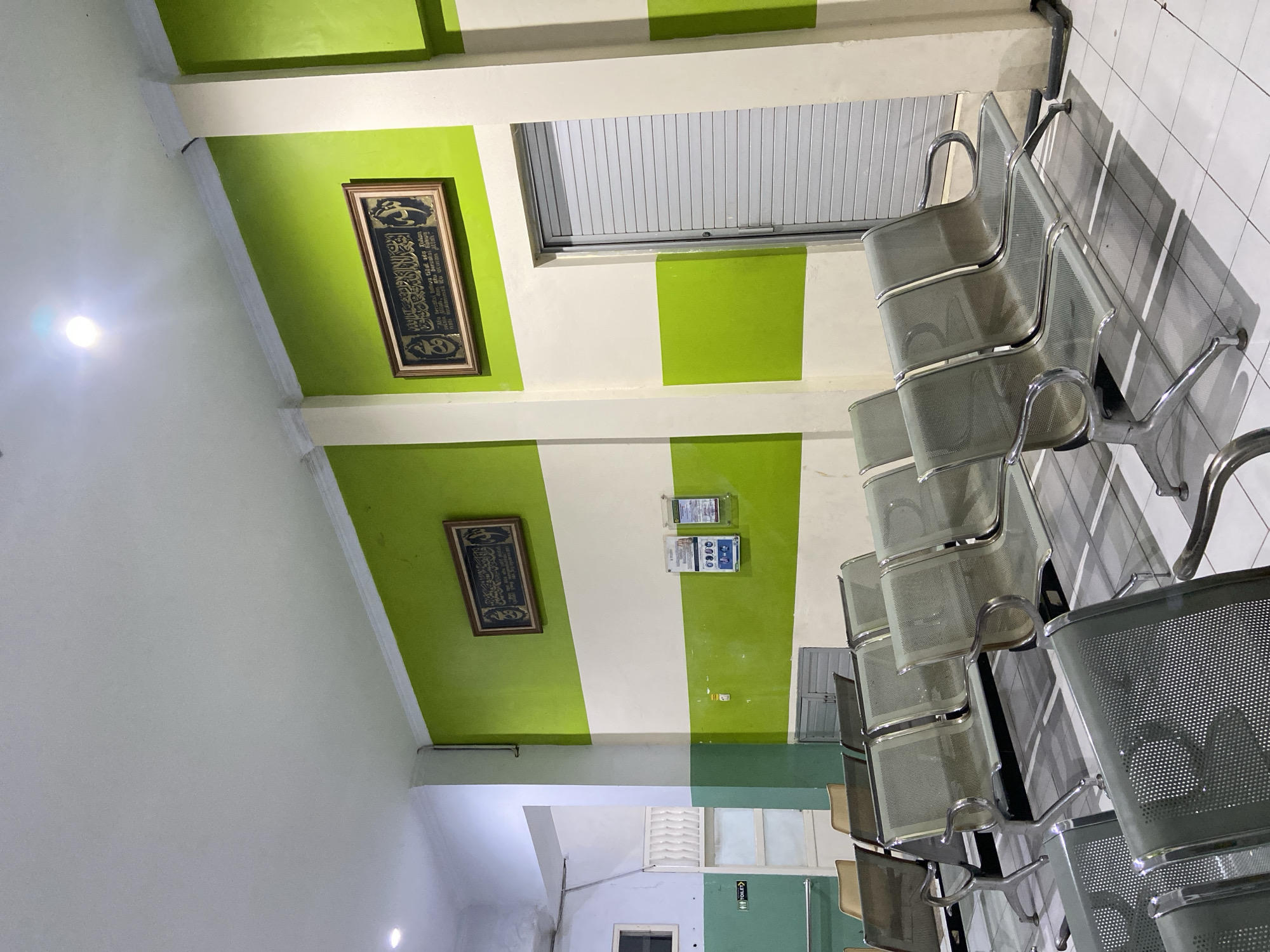 This media file shows a waiting area, likely in a public building or institution, characterized by its green and white striped walls and tiled floor. The scene is dominated by rows of metal chairs, suggesting a place designed to accommodate multiple people waiting for an appointment or service. The chairs are modern, with a mesh-like material for the seat and back, and chrome accents.
On the walls, there are two framed pieces of calligraphy, likely religious texts, adding a cultural or spiritual element to the environment. Above the chairs, a horizontal window with vertical blinds is visible. There are also some signs on the wall, one of which appears to display information with icons, possibly related to health or safety guidelines. Another sign clearly indicates "TOILET" with an icon.
The lighting suggests it is daytime, with a prominent light fixture visible on the white ceiling. The overall impression is one of a clean, functional, and organized space, typical of a waiting room. The image is taken from a low angle, emphasizing the rows of chairs and the striped pattern of the walls. The scene appears quiet and empty, with no people present, implying the waiting area is currently unoccupied. The location context provided is Margoyoso, Indonesia.