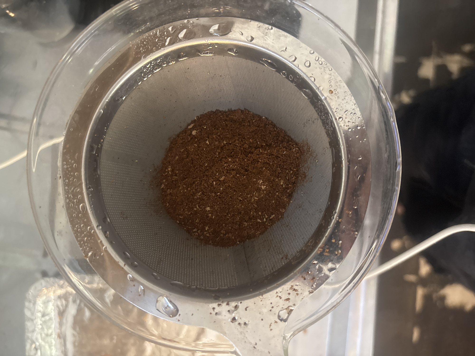 An overhead, close-up shot captures the interior of a clear, possibly glass or plastic, pour-over coffee brewer or tea infuser. Inside the vessel is a stainless steel mesh filter, which holds a substantial mound of dry, coarse-ground coffee or tea leaves. The grounds are a rich brown color with some lighter flecks. Numerous small water droplets are visible on the inner and outer surfaces of the clear vessel, as well as on the metal rim of the filter, suggesting it might have been recently rinsed or is being prepared for brewing. The background is blurred, hinting at an indoor setting, possibly a kitchen counter, and a white cable can be seen on the right side. No people are visible, and there is no discernible text. The image provides a close look at the initial stage of preparing a beverage. The context given for the image is Buenos Aires, Argentina.