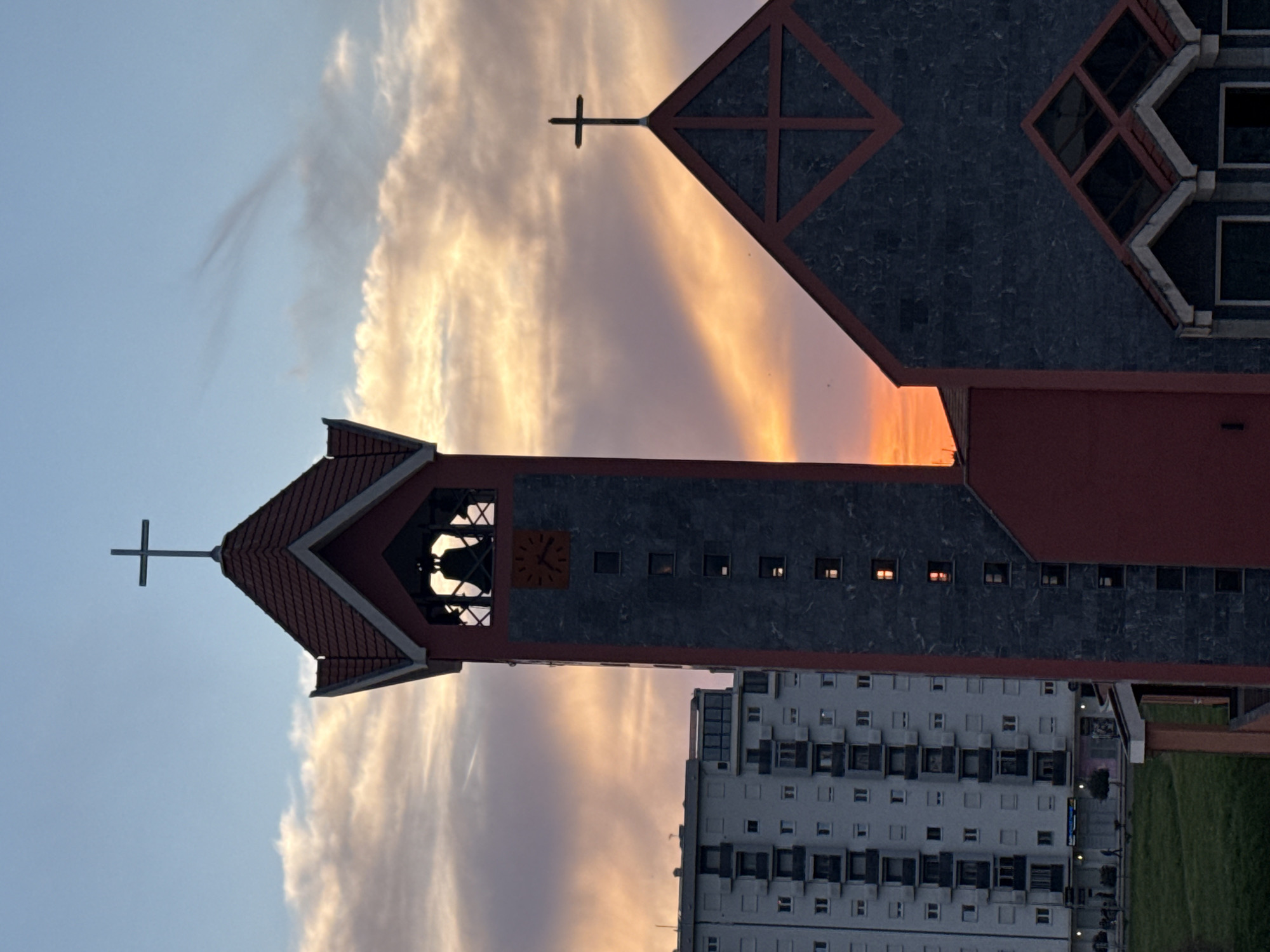 A low-angle, portrait-oriented outdoor shot depicts the Saint Peter the Apostle Church in Bar, Montenegro, against a backdrop of a partly cloudy sky tinged with orange and purple hues. The church features two prominent structures: a tower on the left, topped with a cross, and a larger, more complex structure on the right, also adorned with a cross and unique window designs. A clock face is visible on the tower. Adjacent to the church is a tall, multi-story residential building. The overall ambiance suggests twilight or sunset.