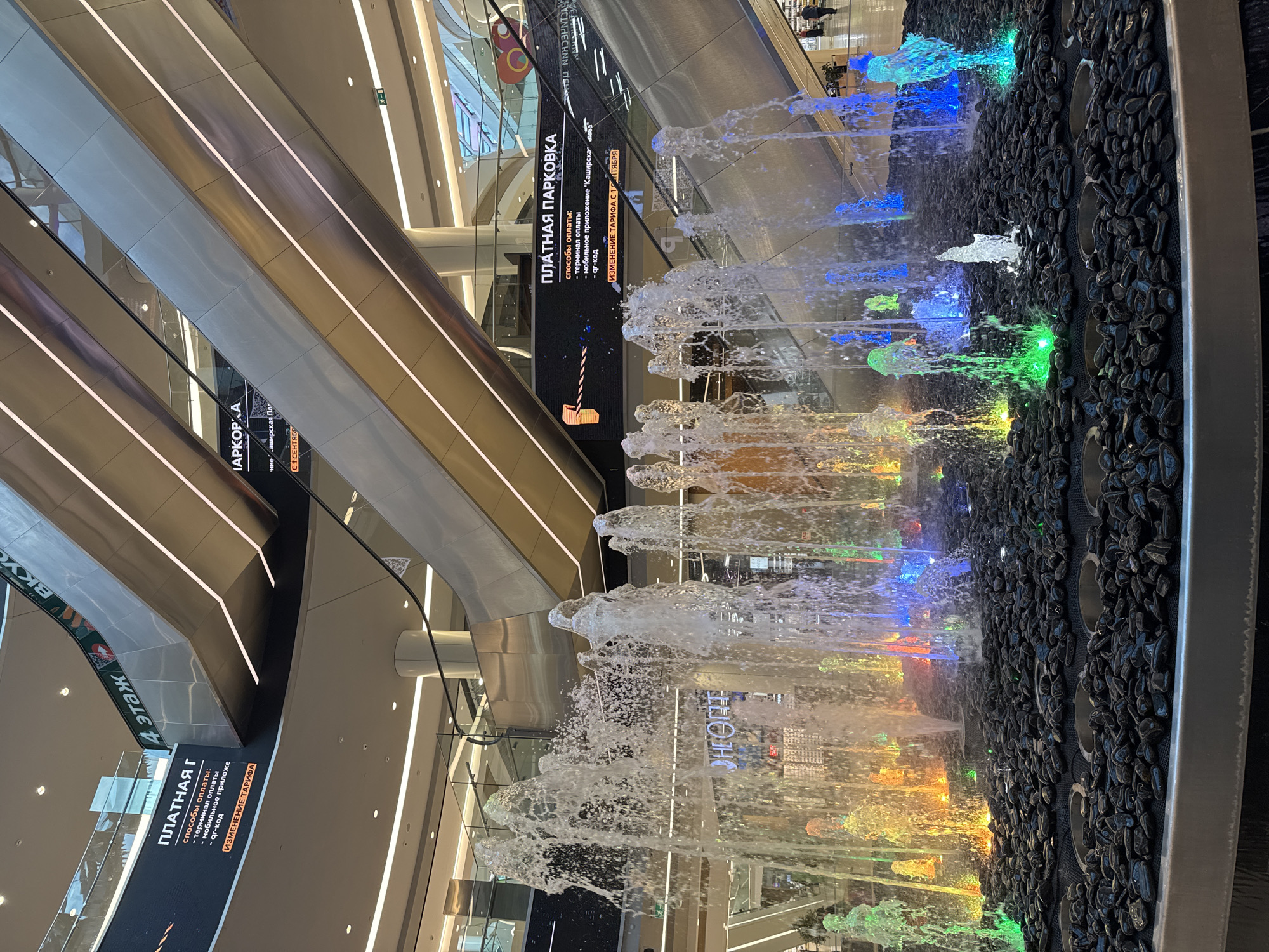 The image depicts an indoor fountain in a modern shopping mall. The fountain features multiple water jets illuminated by colorful lights, creating a vibrant display. The background shows escalators and architectural elements of the mall, including signage. One sign in Russian indicates "ПЛАТНАЯ ПАРКОВКА" (Paid Parking). Another sign visible in the background reads "4 ЭТАЖ" (4th Floor). The environment appears clean and well-lit, typical of a large retail space. No people are prominently featured in the foreground, though a few figures can be seen in the distance on an upper level. The fountain is surrounded by dark-colored decorative stones. The overall scene suggests a bustling commercial center.