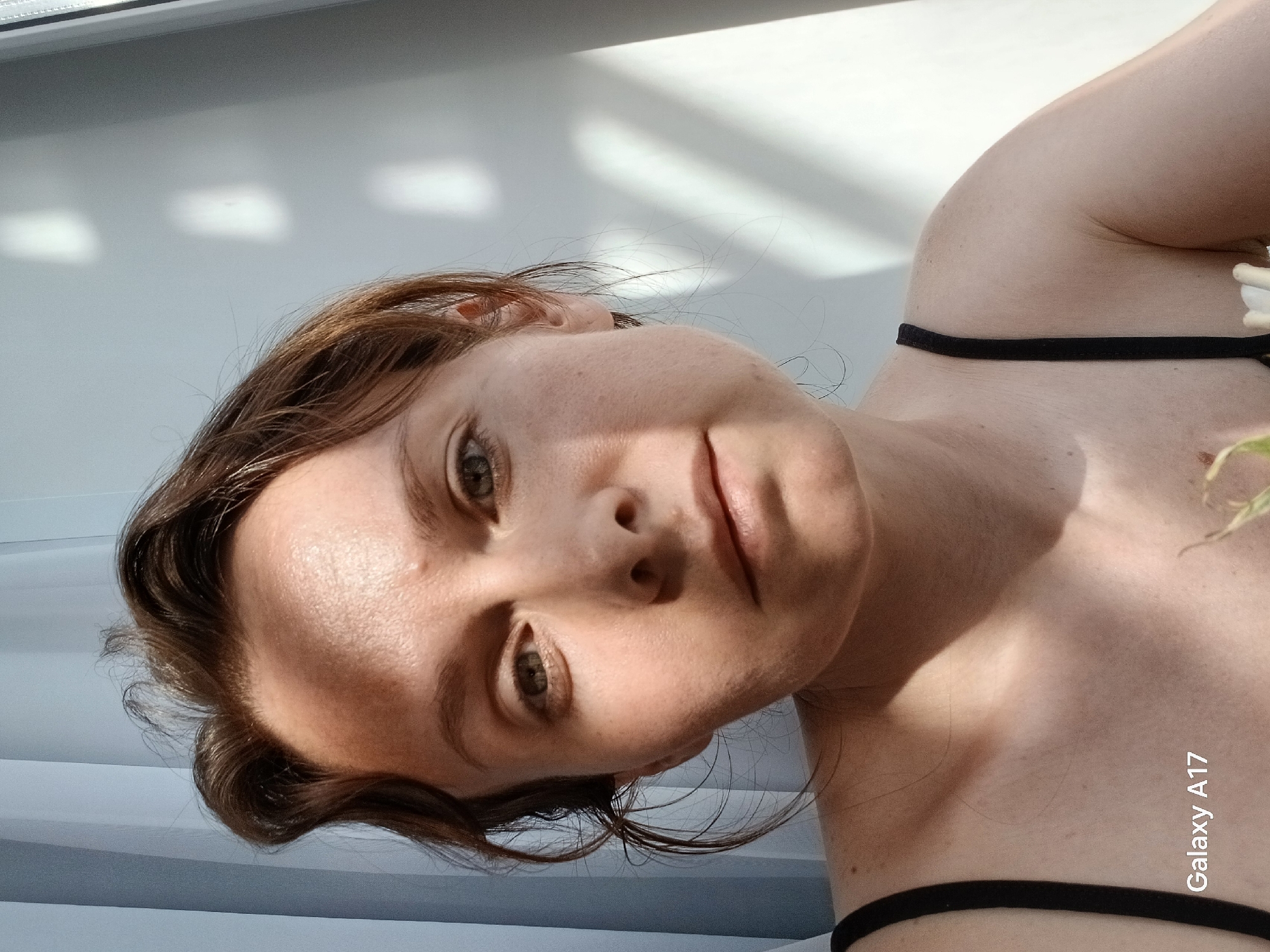 A close-up, slightly angled self-portrait of a fair-skinned woman with brown hair and green eyes. She is wearing a black tank top. The background appears to be an interior space with a window on the left, casting sunlight and shadows on the wall behind her. The location, Yekaterinburg, Russia, cannot be determined from the visual information provided.