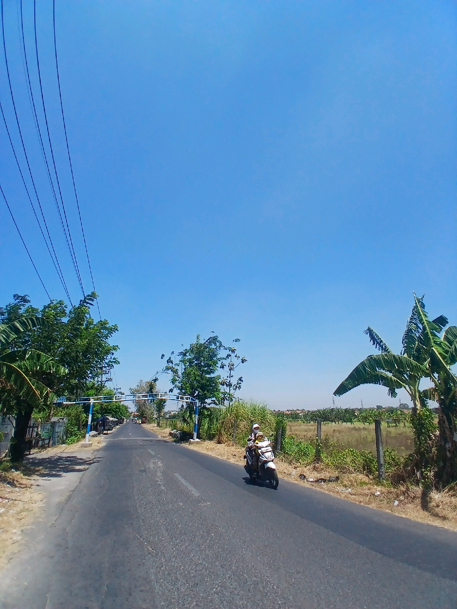 The image depicts a rural road in Surabaya, Indonesia, on a clear, sunny day. A single motorcycle with two occupants, a driver and a passenger, is traveling away from the camera on the asphalt road. The driver is wearing a helmet and a white shirt, while the passenger is wearing a yellow shirt. The road is flanked by dry vegetation and trees, including banana trees. Overhead, power lines stretch across the bright blue sky. In the distance, a white and blue archway spans the road, possibly indicating an entrance to a village or area. The scene is consistent with a typical rural landscape in Surabaya.