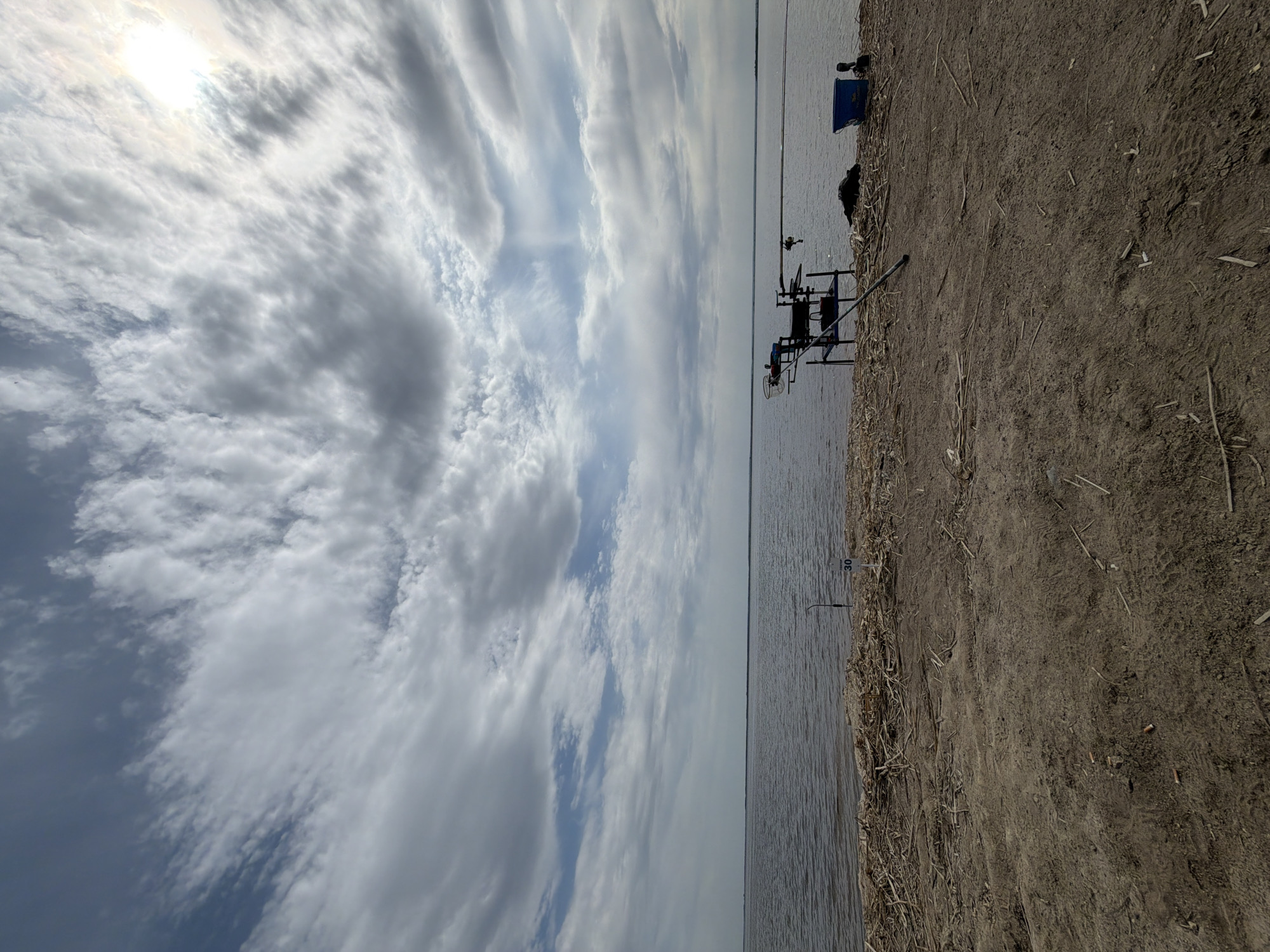 The image shows a lakeside view on what appears to be a cloudy day near Krasnyi Bor, Russia. A body of water (possibly a lake or river) occupies the middle ground, with a shoreline on the right. On the shoreline, there's a fishing setup which includes fishing rods, a fishing rod stand, and a blue container. The foreground shows dry, earthen ground with some dried vegetation. In the sky, clouds dominate the view. The sun is visible behind some clouds.