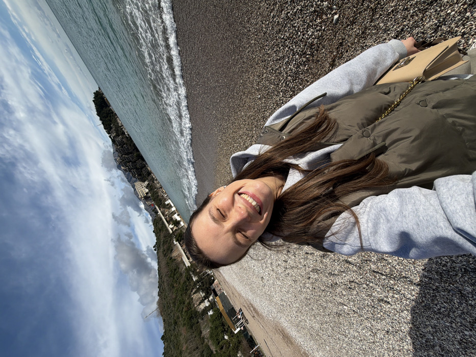 An outdoor, eye-level selfie captures a young adult female on a pebble beach in Boreti, Montenegro. The female subject, positioned in the lower-center of the frame, has long, straight, dark brown hair and is smiling broadly with her eyes closed. She wears a light grey hooded sweatshirt under a dark olive green puffer vest. A light tan shoulder bag with a gold chain strap is visible on her right side. The beach consists of small, light-colored pebbles. To the right, the light blue-green sea exhibits small breaking waves at the shore. The background features a steep, tree-covered hillside with several white buildings, likely hotels or resorts, visible along the slope and near the waterline. Distant mountains rise further behind. The sky overhead shows patches of blue sky interspersed with white and grey clouds. The scene is brightly illuminated by natural sunlight.