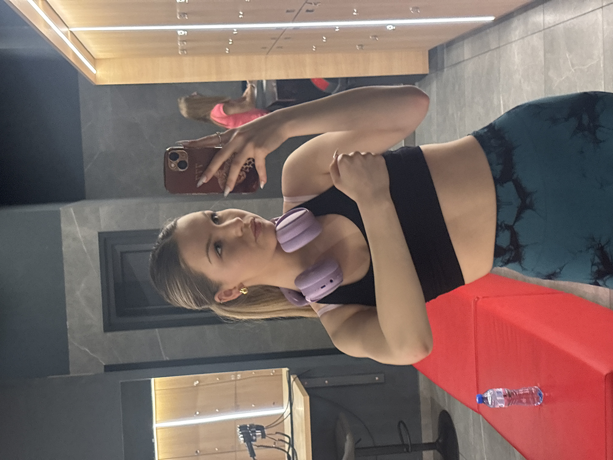 The image captures a young woman taking a mirror selfie indoors, likely within a gym or changing room setting in Plovdiv, Bulgaria. She is positioned with her body slightly angled, holding a mobile phone with a red-brown case in her right hand, extending it towards the mirror to capture her reflection. Her long, light-colored hair is styled back, and she is wearing a black sports bra and teal-patterned athletic leggings. A pair of light purple over-ear headphones rests around her neck, and she has a small gold stud earring in her right ear. Her expression is neutral, and her gaze is directed slightly off-camera.
In the background, a row of light wooden lockers with horizontal strip lighting at the top is visible, reflecting in the mirror. Faintly visible in the locker reflection is another individual with dark hair, wearing a bright pink top. To the woman's left, a red padded bench is present, upon which a clear plastic water bottle with a blue cap rests. The walls feature grey tiling, contributing to a clean and modern aesthetic. The specific time of day is not indicated due to the indoor lighting. The phone case has some faint, stylized text that appears to be "TTT" or similar, and her nails are long and polished.