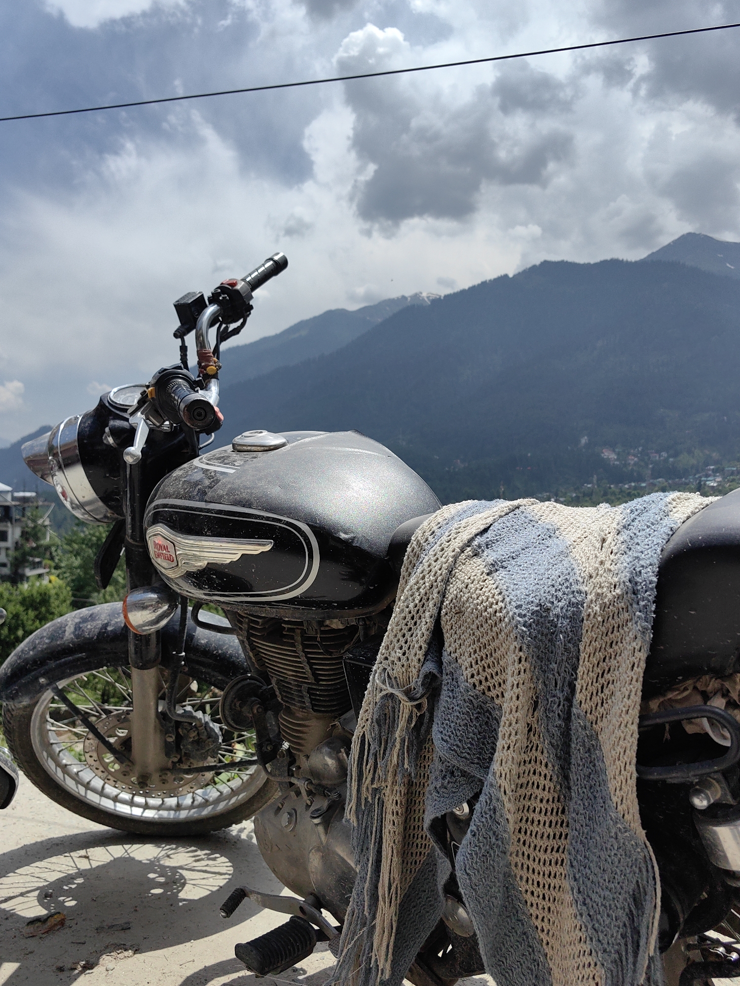 A black Royal Enfield motorcycle, appearing well-used with visible dust and grime on its fuel tank, engine, and front fender, is parked on a dirt or gravel path under a partly cloudy sky. The motorcycle features a chrome headlight, spoked wheels, a front disc brake, and the "ROYAL ENFIELD" winged emblem on its fuel tank. A light blue and beige striped, knitted or crocheted blanket is draped over the rear seat and side of the motorcycle. A small orange-red decorative element is affixed near the left handlebar. In the background, dark blue-green, forested mountains rise steeply, with some distant peaks showing traces of snow. A valley containing a scattered settlement with buildings and greenery is visible below the mountain range in the far distance. A thin, dark overhead line traverses the upper sky. Strong shadows, including a clear outline of the motorcycle's front wheel and disc brake, are cast on the ground. The scene is located in the mountainous region of Vashisht, India. No individuals are present.