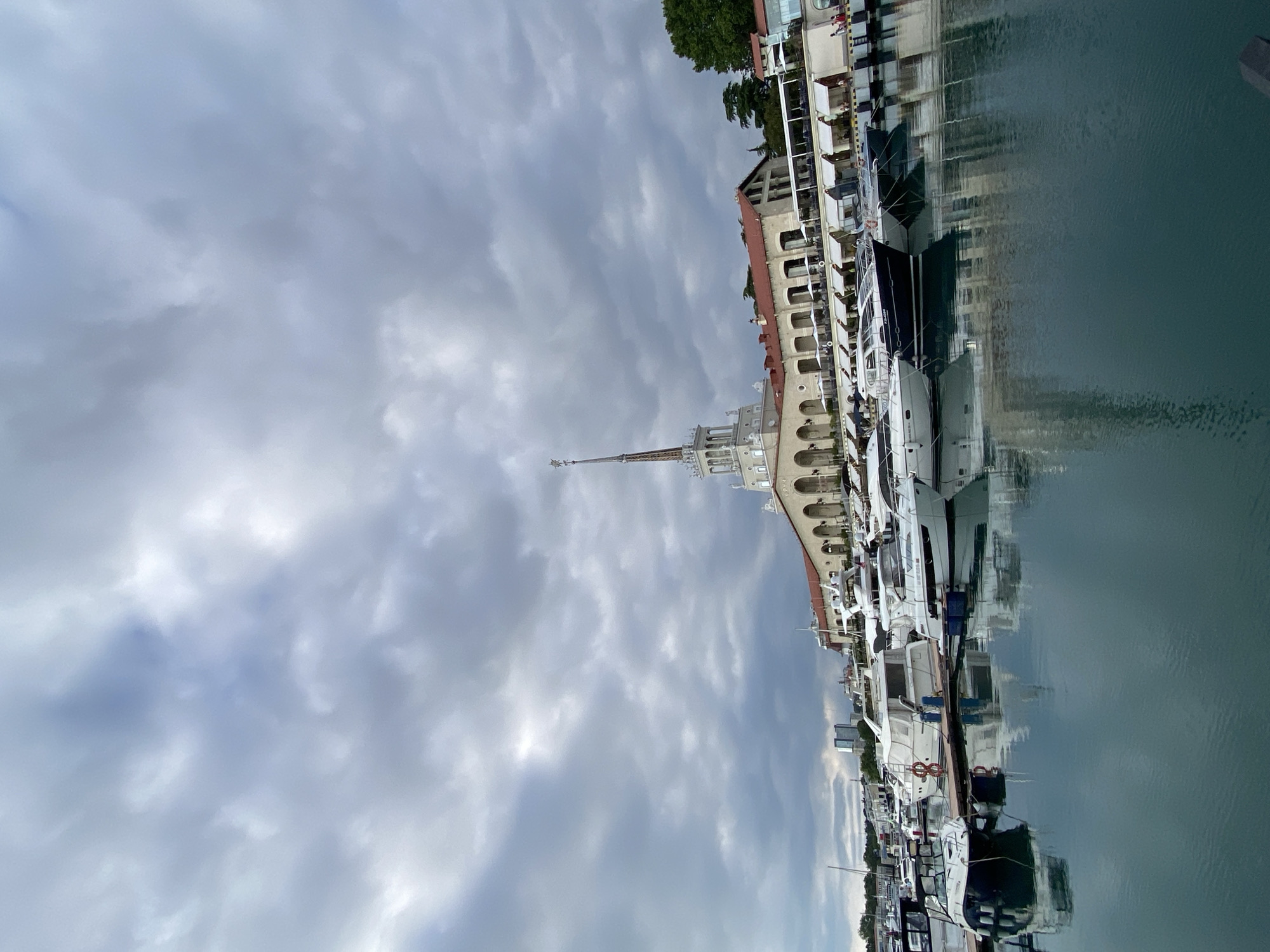 The image depicts a view of a harbor in Sochi, Russia, with several yachts docked along a waterfront promenade. The architecture of the buildings lining the harbor features arched windows and red-tiled roofs, reflecting the classical style often seen in the region. A prominent tower with a spire stands out against the cloudy sky. The water in the harbor is calm, with clear reflections of the boats and buildings.
The weather appears overcast, with a sky filled with a mix of grey and white clouds. This suggests it might be a cloudy day, possibly with the threat of rain, or simply a diffused light condition. The time of day is not explicitly clear due to the cloud cover, but the overall lighting suggests it is daytime.
The scene is a serene harbor, likely a popular tourist area given the prominent architecture and numerous docked vessels. The image captures the stillness of the water and the architecture, creating a sense of peacefulness. No specific activity or event is clearly visible, other than the stationary boats. There are no visible people in the foreground, although there might be individuals present within the buildings or on the yachts, it is difficult to discern.
There is no discernible text visible in the image.