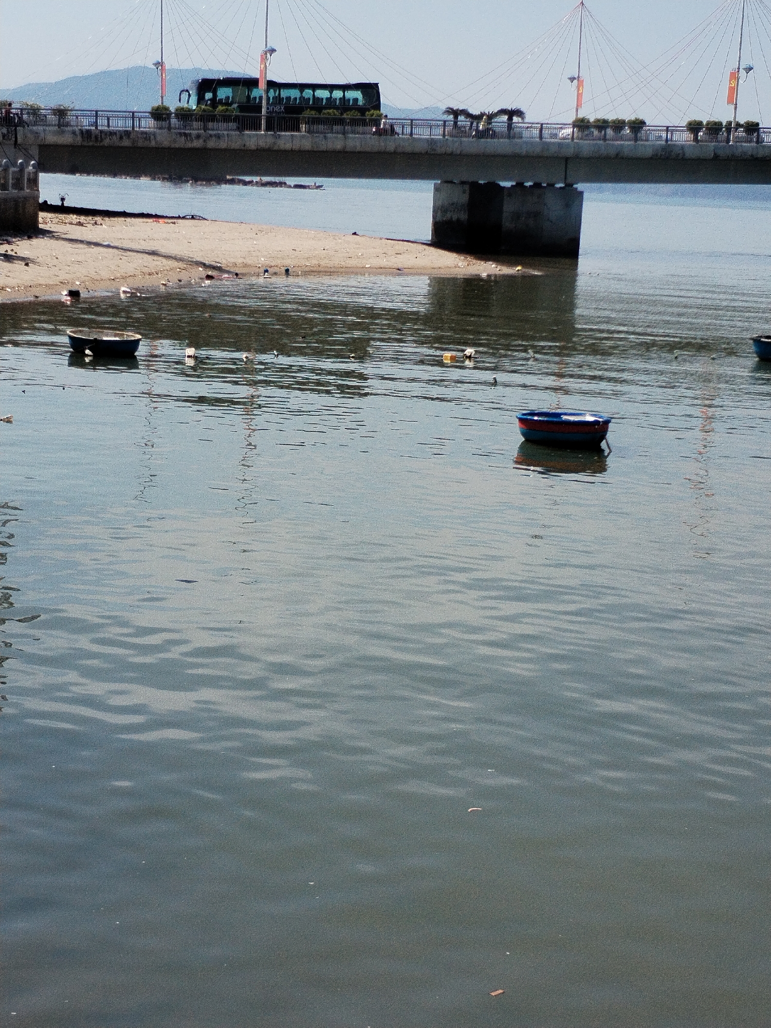 The image shows a section of riverfront, possibly in Bac Nha Trang Ward, Vietnam, as indicated by the location. In the middle ground, there is a body of water, likely the river, with slight ripples and some debris floating on the surface. Two small, round boats are visible in the water. One is dark and relatively distant, while the other is closer and appears to be painted with blue and red colors. In the background, there's a sandy area with litter scattered across it. Behind the sandy area, there's a bridge supported by concrete pillars. A bus is crossing the bridge. The overall impression is one of a somewhat polluted environment.
