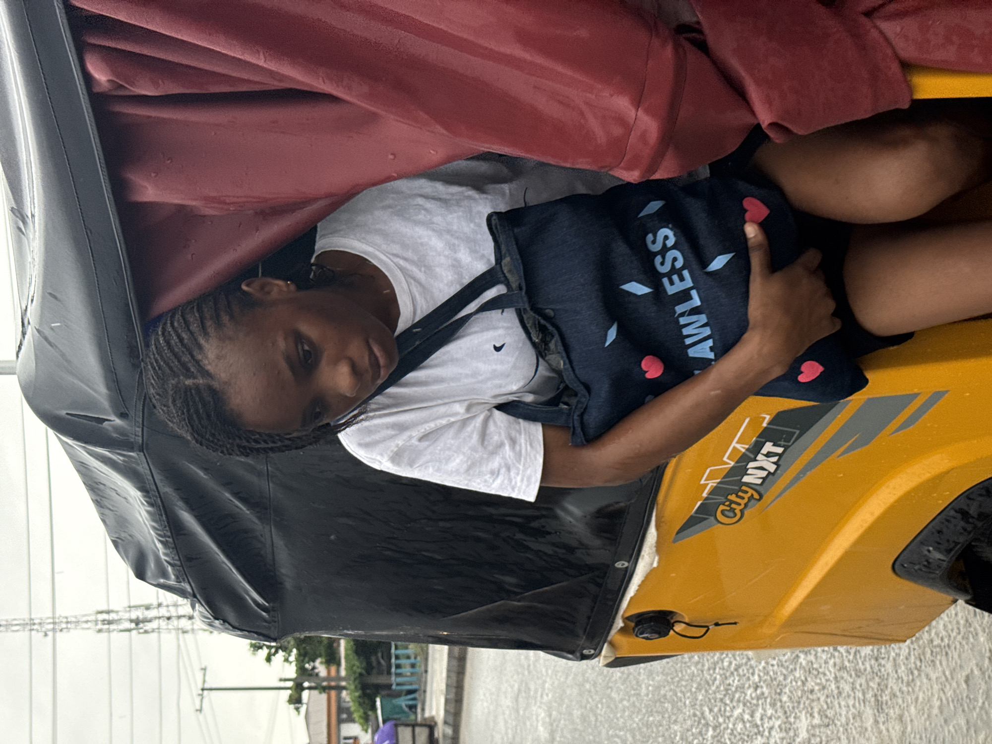 A young woman rides inside a yellow "City NXT" tuk-tuk in Ado, Nigeria. The tuk-tuk has a black canvas roof, a red vinyl sheet, and is driving on a flooded street. The woman wears a white t-shirt and holds a dark blue bag that has the word "AWLESS" printed on it, as well as small red hearts. The background shows street lights and buildings partially obscured by the heavy rain.