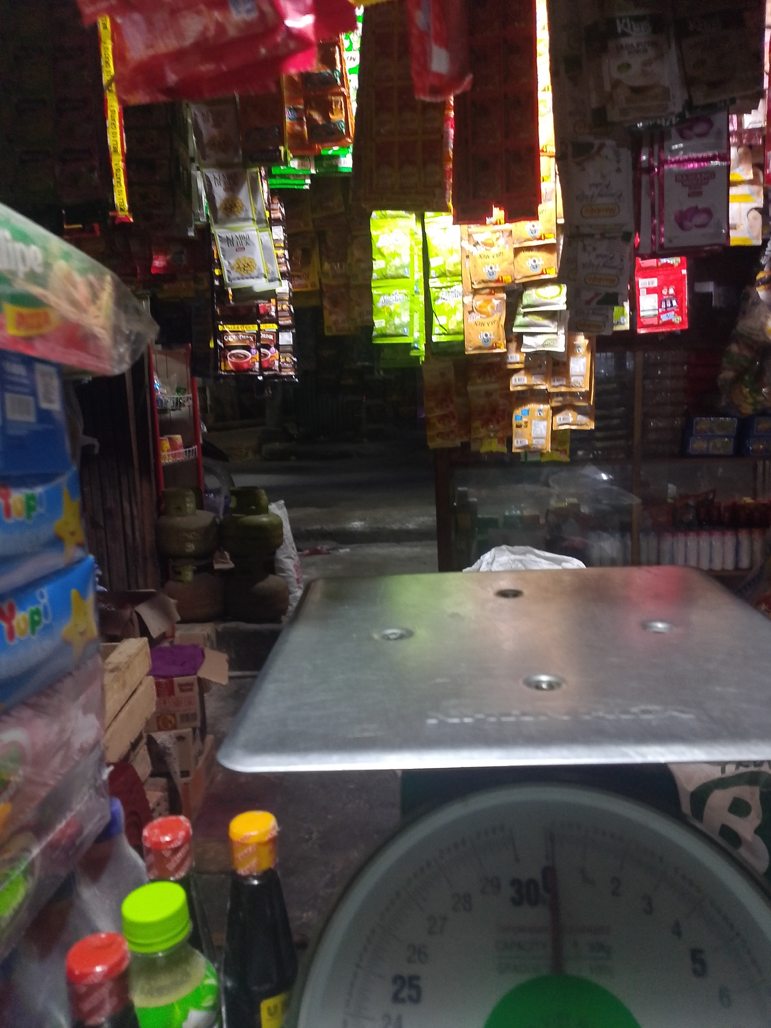 The image depicts a cluttered street-level shop interior, likely in Palembang, Indonesia. The foreground is dominated by a large, metallic weighing scale with a dial marked with numbers and measurement units. Several bottles with colorful caps are visible on the surface of the scale and to its left, including one with a bright green cap and another with a yellow cap. In the background, shelves are packed with various packaged goods, predominantly food items and beverages, displaying a wide array of colors and brands. Some items are hanging from the ceiling, further adding to the sense of a densely stocked retail space. Two large gas cylinders are partially visible near the back, suggesting the shop might also sell household essentials. The lighting is dim, with some areas illuminated by overhead or ambient light, creating a somewhat shadowy atmosphere. The overall impression is that of a small, traditional Indonesian convenience store or "warung." The presence of gas cylinders and the dense display of packaged goods are common characteristics of such establishments in Palembang and other Indonesian cities.