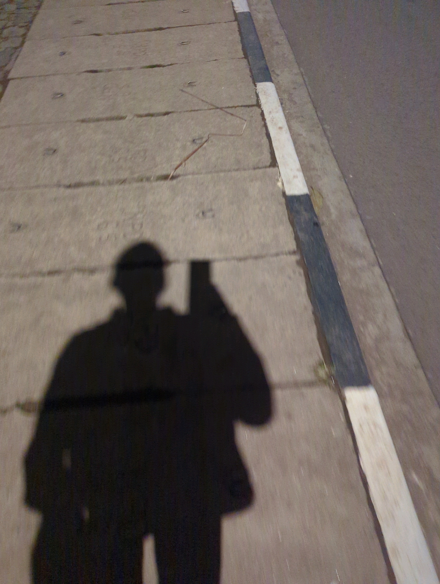 The image is a low-angle, shadow selfie taken on a sidewalk next to a road in Musanze District, Rwanda. The shadow of a person holding a phone is cast on the concrete sidewalk. The sidewalk is made of rectangular concrete slabs with visible seams and some small circular metal fixtures embedded in them. A dark and white striped curb separates the sidewalk from the asphalt road. The lighting suggests it may be dusk or night, with artificial light illuminating the scene.