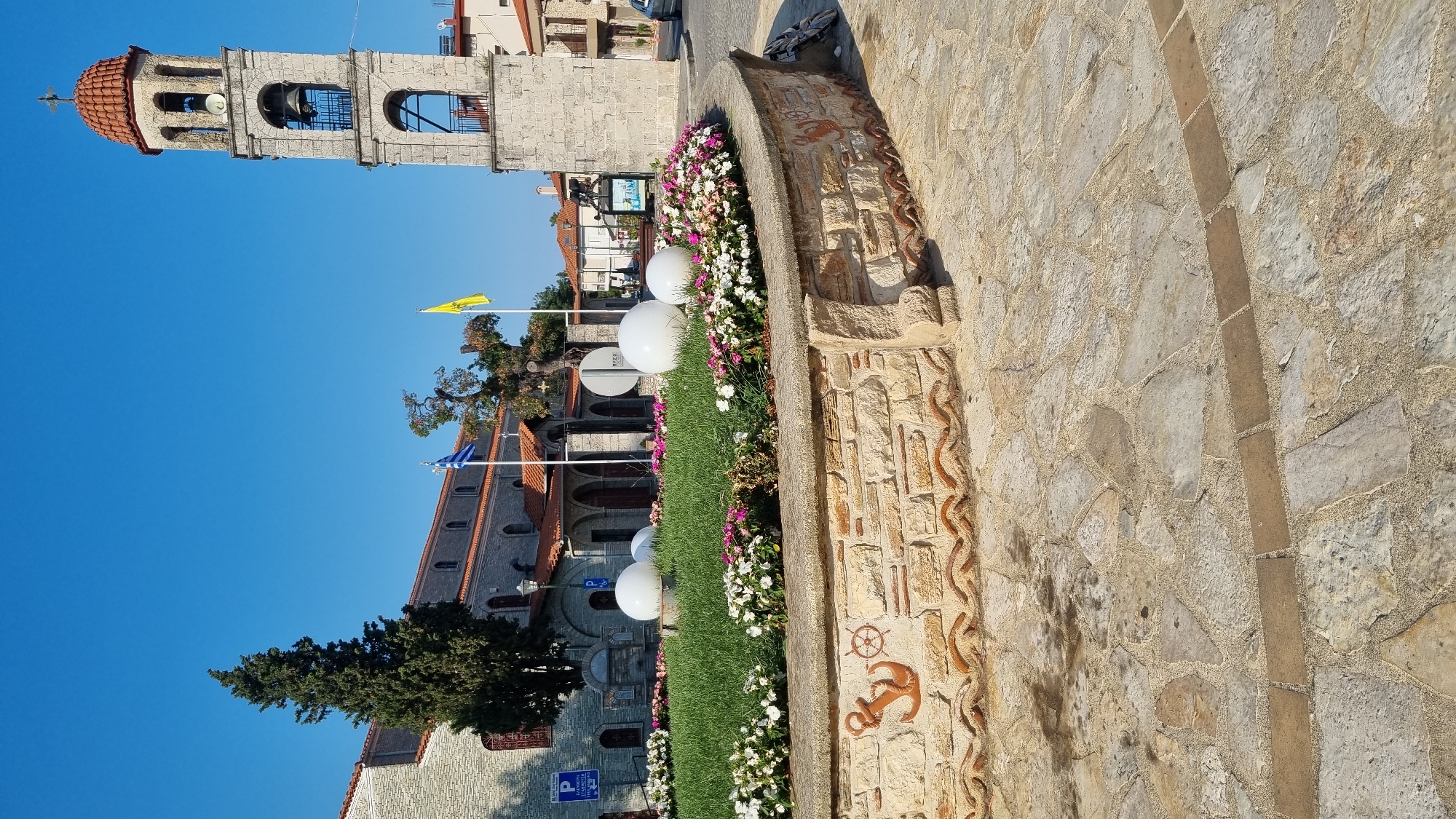 This image captures a public space in Litochoro, Greece, featuring a stone-paved area in the foreground with a circular flowerbed at its center. The flowerbed is bordered by a low, multi-textured stone wall adorned with decorative elements, including an anchor and a ship's wheel motif. Inside the flowerbed, vibrant green grass is interspersed with blooming flowers in shades of pink and white, and two large, white spherical objects are placed strategically.
In the background, a multi-tiered bell tower with a red tiled roof stands prominently. Adjacent to the tower is a stone building with an arched entrance, likely a church, indicated by a cross at the apex of its roof. The Greek flag and a yellow flag are displayed near the buildings. Several trees, including a tall cypress tree, are visible, adding to the greenery of the scene. The sky above is a clear, bright blue, suggesting fair weather. A street with parked cars is visible to the right, and a signpost with Greek text is present near the left side of the frame, indicating a parking area. The overall impression is of a well-maintained public square with architectural and natural elements, typical of a Greek town.