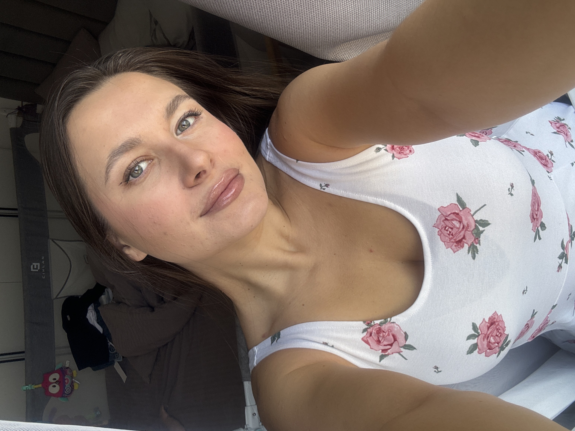 A woman is seen in a selfie. She has brown hair, fair skin, and is wearing a white tank top with a pink rose pattern. The background includes part of a room with bedding, a gray chair, and some miscellaneous items. There is no information to ascertain the location is in the city of Odintsovo, Russia.