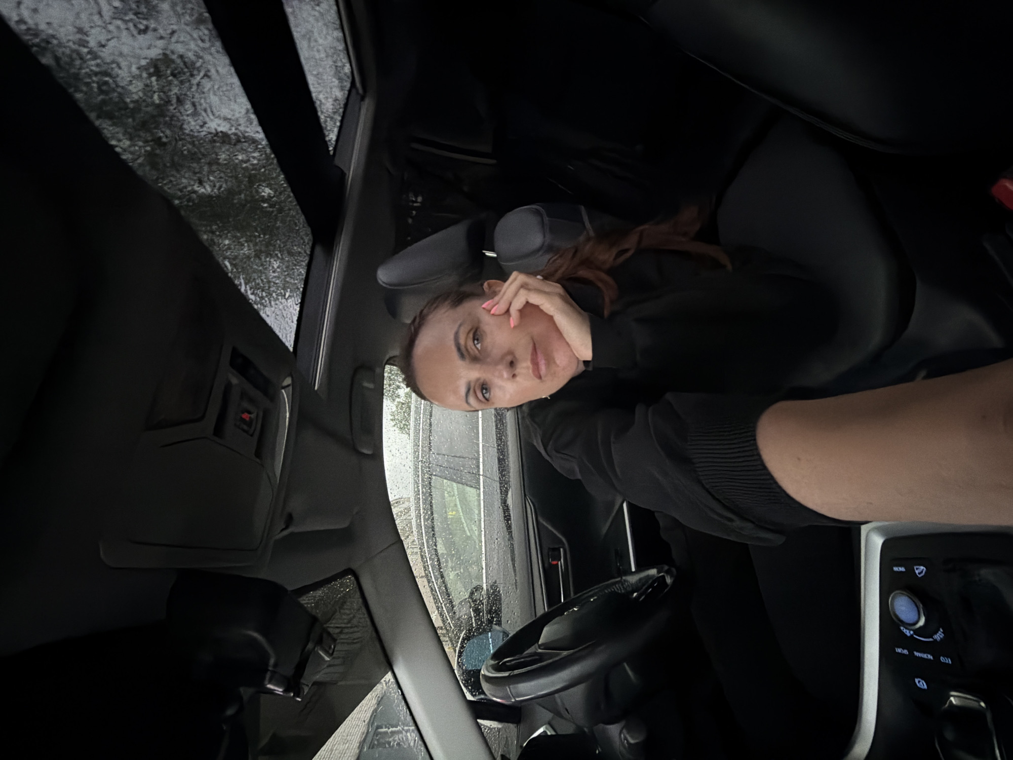 The image captures a woman inside a car, with the camera positioned from an unusual, sideways angle. The woman is looking directly at the camera, her right hand resting on her cheek. She has dark hair, which appears to be pulled back loosely, and she is wearing a dark-colored hooded sweatshirt. Her fingernails are painted a bright pink.
The setting is clearly the interior of a vehicle. We can see parts of the car's dashboard, including the steering wheel and gear shift, as well as the car's interior lights and roof lining. Outside the car windows, the weather appears to be rainy, with water streaking down the glass and a blurry, wet landscape visible. The overall lighting suggests it might be daytime, but the overcast and rainy conditions create a subdued atmosphere. The image is oriented sideways, with the top of the image pointing to the left. No visible text is present in the image.