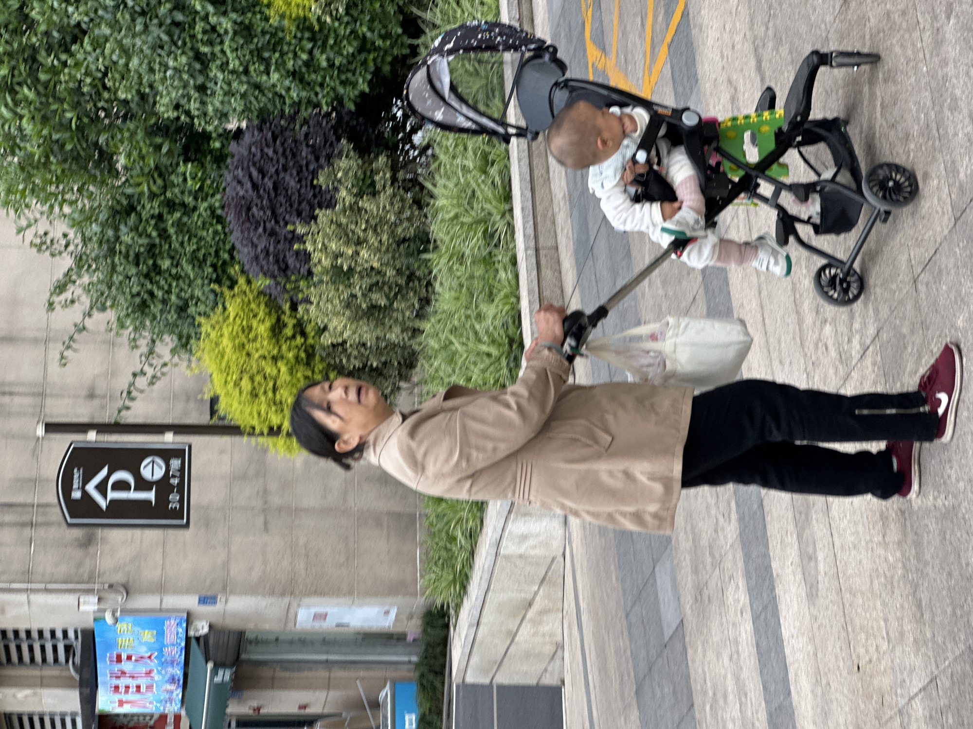 An adult woman is depicted pushing a baby in a stroller along a paved sidewalk in what appears to be an urban environment in Jiangjin, China. The image is taken at eye-level, showing the woman from the waist up and the baby in the stroller.
The woman, with dark hair pulled back, wears a light brown or tan jacket over black pants and maroon sneakers with white soles. Her face is turned slightly upwards, and she has an open-mouthed expression, suggesting she might be talking or laughing. She is holding onto the handle of the stroller, from which a white plastic bag is also hanging.
Seated in the stroller is a baby or toddler, wearing a white long-sleeved top, light-colored pants, and white shoes. The child has light brown hair and is looking forward, secured by the stroller's harness. The stroller itself is black with a patterned canopy and green accents on the seat. Yellow lines are visible on the ground near the stroller, possibly indicating a traffic or parking area.
To the left of the woman, a black street sign with white text is mounted on a concrete wall. The sign displays a stylized "P" inside an inverted triangle, a symbol resembling a simplified vehicle, and Chinese characters "310-47建". Below this sign, part of a blue poster or advertisement with red and white Chinese characters is visible, though the text is not clearly legible.
The background features lush greenery, including dark green bushes, lighter green foliage, and some darker purple or black shrubs, suggesting a planted area or park beside the sidewalk. The lighting appears even, indicating a daytime scene, possibly under an overcast sky, as there are no harsh shadows. The overall scene captures a casual moment of a caregiver and child outdoors.