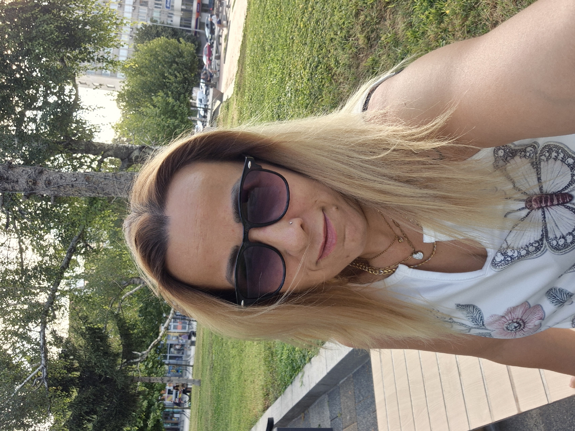 A woman with blonde hair and sunglasses is taking a selfie outdoors. She is wearing a white tank top with a floral and butterfly pattern and two necklaces. The background shows a grassy park area with trees and buildings in the distance. The sun appears to be setting, casting a warm glow on the scene.