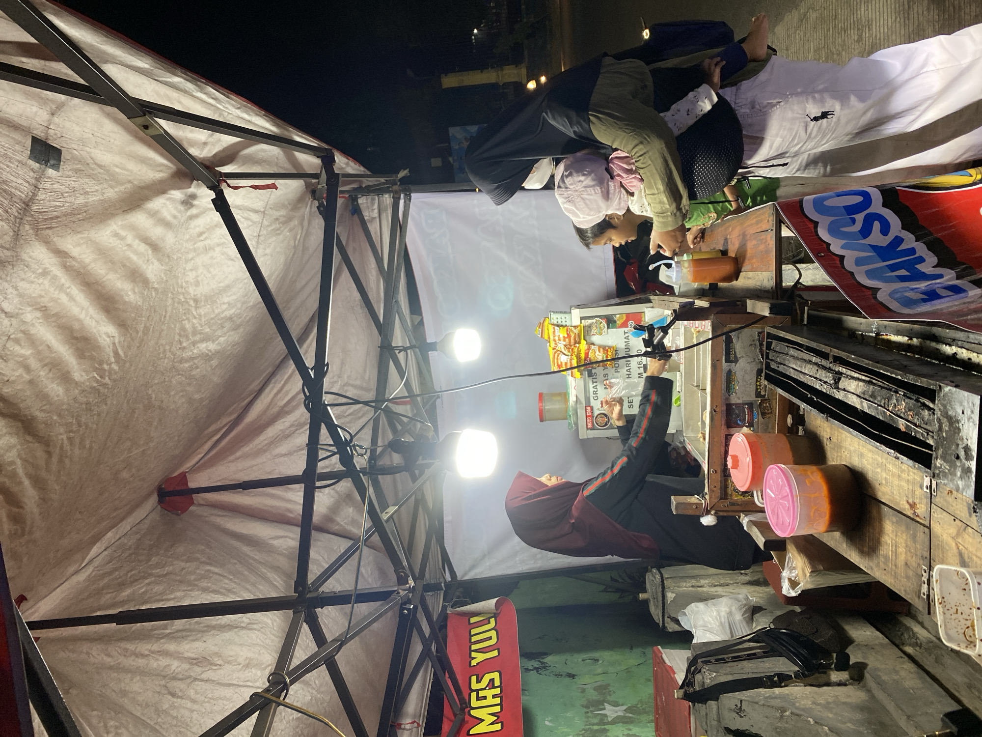 The image depicts an outdoor food stall at night in the city of Kasihan, Indonesia. The stall, likely a mobile cart or temporary setup, is covered by a large white tarp supported by a metal frame. It is illuminated by artificial lights.
In the foreground, a wooden counter displays various items, including large plastic containers filled with sauces or liquids, and what appears to be a grilling or heating apparatus. A black bag is visible on the left, hanging from a concrete structure. A red and white sign with blue lettering that reads "BAKSO" (a type of Indonesian meatball soup) is prominently displayed at the bottom right.
There are three people visible. A woman wearing a dark hijab and a black jacket with red stripes is standing behind the counter, interacting with a customer. She appears to be serving or preparing food. To the right of the counter, a woman wearing a black hijab and a green jacket is holding a baby in a carrier. A young child, possibly a boy, is standing next to her, looking towards the stall.
The background shows a dimly lit street with some buildings and a lamppost visible, suggesting an urban or semi-urban environment. The overall scene captures a moment of commerce and daily life at a street food vendor in Indonesia.