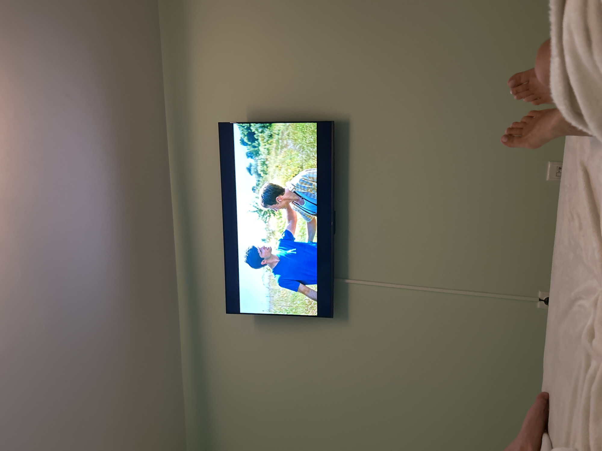 The image depicts a flat-screen television mounted on a light green wall. The television displays a scene with two young men outdoors, with one wearing a blue t-shirt and the other a checkered shirt. Below the television, a white cable runs down the wall and connects to an electrical outlet. In the foreground, a person's feet are visible, covered by a white blanket, suggesting they are lying in bed. The ceiling above is a neutral color. The scene appears to be indoors, in a bedroom or living area. There is no discernible information in the image that places the scene within the city of Burgas, Bulgaria.