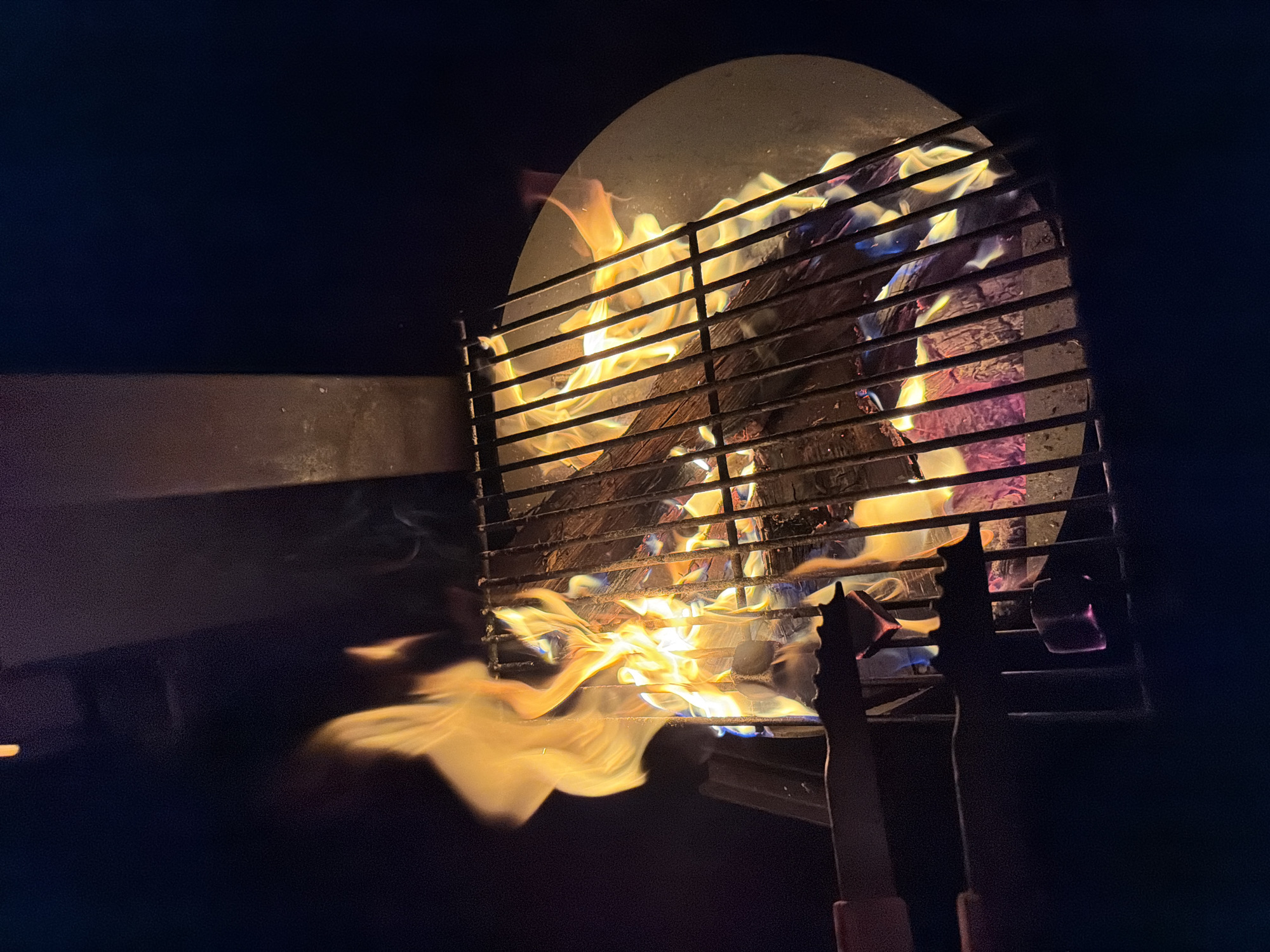 An outdoor fire pit is visible from a top-down perspective, with a metal grate covering the fire. Flames erupt from burning logs within the pit, casting a warm orange glow that illuminates the surrounding area. Smoke drifts upwards from the fire, dissipating into the dark night sky. The fire pit appears to be made of metal, with a circular base and a vertical support structure visible at the top of the frame. Two sets of tongs are positioned over the grate, suggesting an intention to cook food over the fire. The scene is set against a dark background, likely indicating nighttime or a dimly lit environment. There are no visible people in the image.
The specific location in Bayreuth, Germany, cannot be determined from the provided image alone.