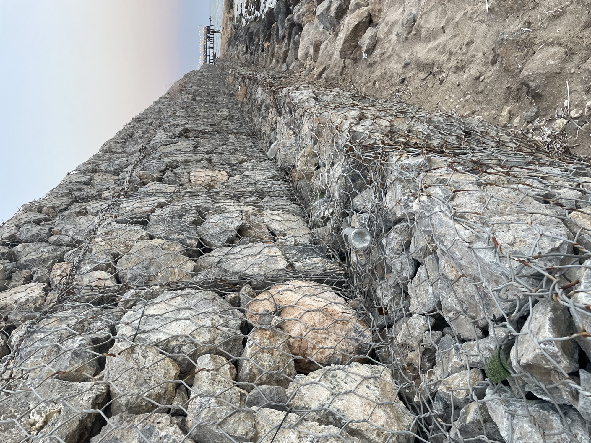 The image shows a gabion wall constructed from rocks and wire mesh along a coastline. The wall extends diagonally from the foreground to the background, where it meets the sea. A pier with lights is visible in the distance, as well as a person walking on the shore. The sky is clear with a gradient of colors, suggesting either sunrise or sunset. Some debris, including a plastic bottle, is present within the gabion wall and on the sandy shore. The area appears to be a beach with scattered rocks. The specific location is the coast of Trikomo, Northern Cyprus.
