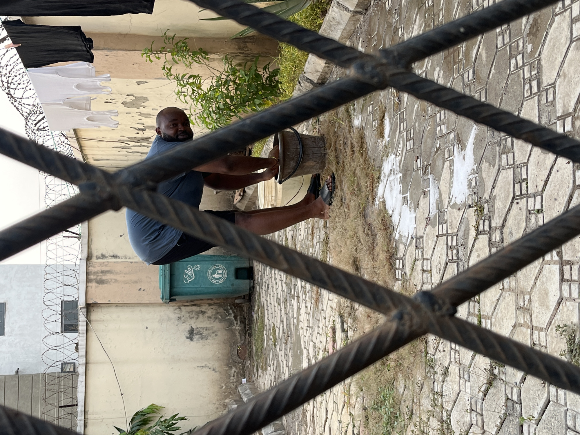 This image, captured in Dushepe, Nigeria, portrays a man engaged in outdoor domestic work, viewed through the prominent, dark, twisted bars of a metal grate or fence. The scene is presented with the man vertically oriented, suggesting a 90-degree rotation from the original capture.
The primary subject is a dark-skinned man with a shaved head and a beard, wearing a dark blue t-shirt and dark shorts. He is barefoot, with a black sandal visible near his left foot. He is focused on a task, with both hands submerged inside a dark, aged bucket resting on the paved ground in front of him, suggesting he is washing or soaking items. White, foamy residue, likely from soap or detergent, is visible on the ground around the bucket.
In the background to the man's left, several white and black sleeveless shirts are hanging on a clothesline, indicating laundry is taking place. Beyond the clothesline, a wall or fence topped with multiple strands of barbed wire runs horizontally. Further back, weathered building walls in shades of light tan and off-white are visible, along with some green foliage.
To the man's right and slightly behind him, a green plastic waste bin with a lid is visible. Text on the bin includes "KEEP CLEAN" and a logo depicting a hand dropping an item into a bin. The ground is paved with patterned concrete or interlocking tiles, interspersed with patches of dry grass and dirt. The time of day appears to be daylight, under natural illumination.