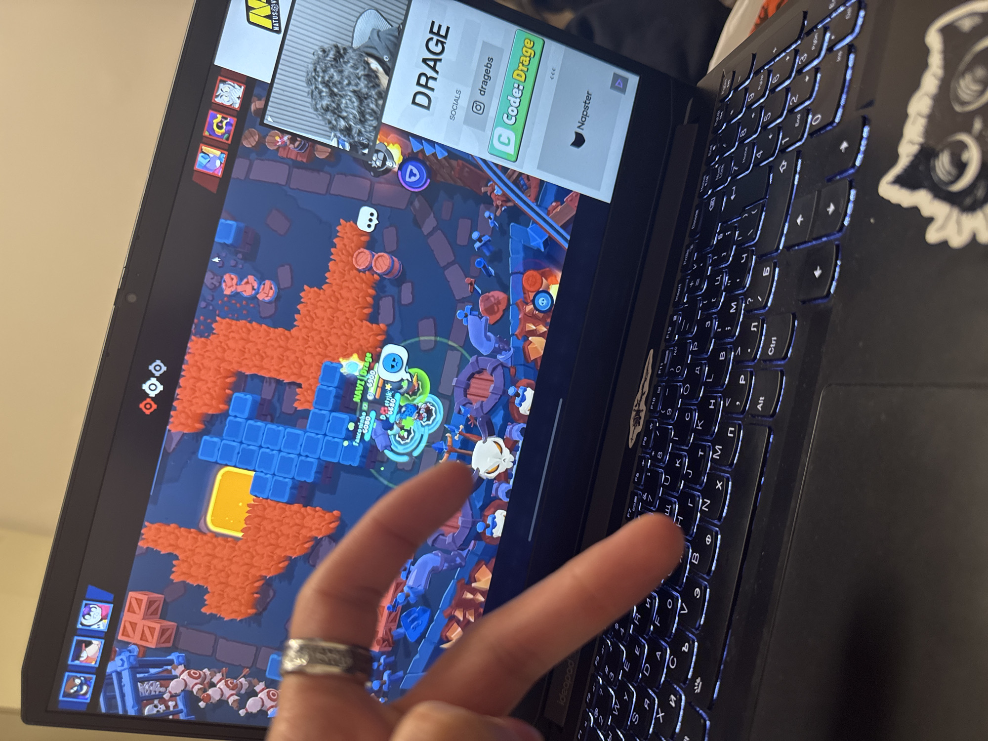 Here's a detailed description of the image:
**Scene:** The image shows a laptop screen displaying the game "Brawl Stars." A hand is partially visible in the foreground, with two fingers extended towards the screen or keyboard.
**Laptop Details:** The laptop has a black exterior with a backlit keyboard that has Cyrillic and Latin characters. There's a sticker of a cartoon owl on the right side of the keyboard. The screen shows the game "Brawl Stars" in progress. There's a smaller window at the top displaying a social media profile or ad for "DRAGE" with the code "Code: Drage".
**Game Details:** The Brawl Stars game is in a battle arena with various characters, obstacles, and terrain.
**Other Details:** A silver ring is visible on the hand.
**Location:** There is not enough information to determine the location in the city of Sofia, Bulgaria.