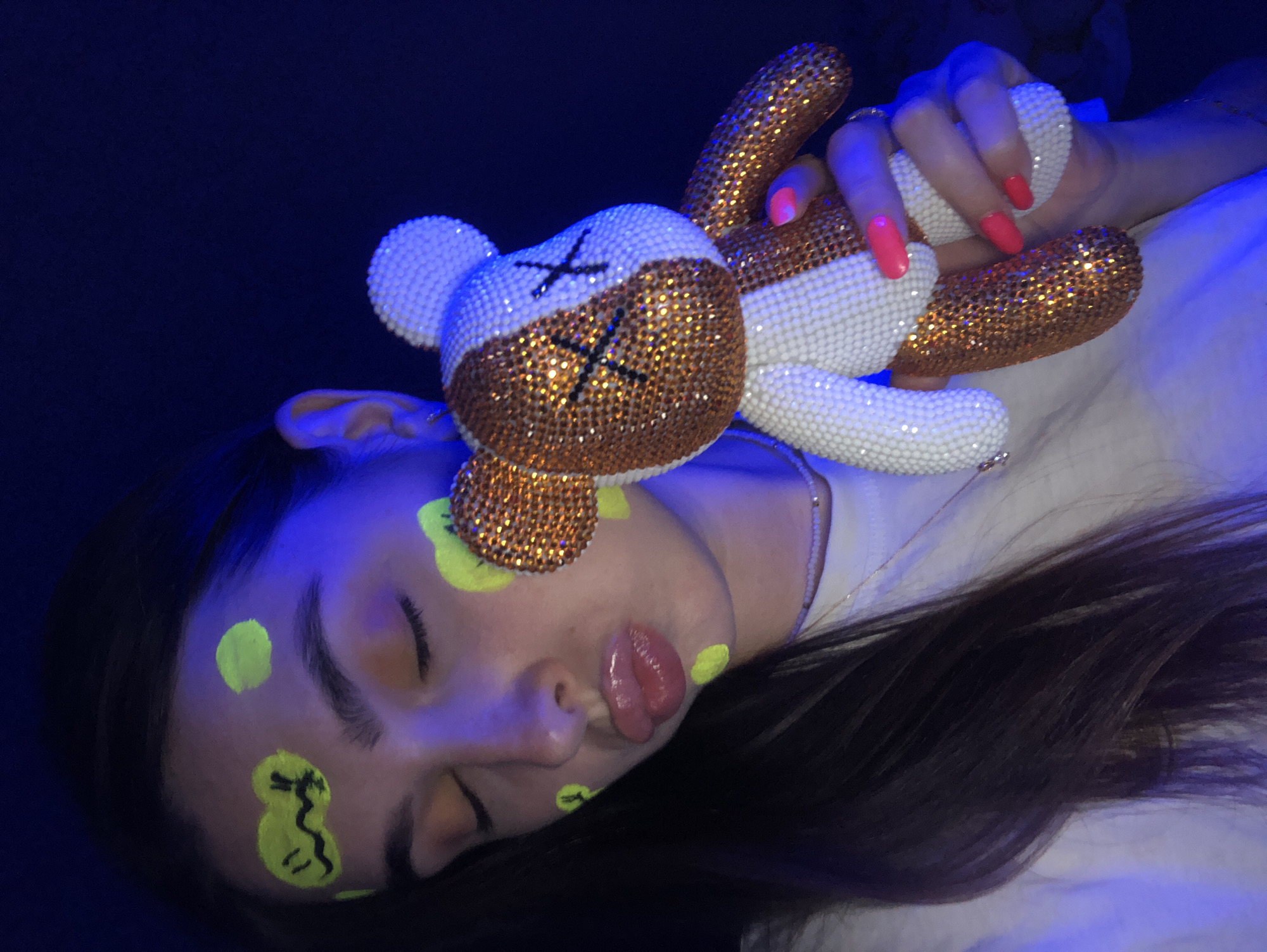 A young woman with dark hair and closed eyes is holding a small stuffed bear. She has neon yellow paint on her face, with shapes resembling a smiley face on her forehead and dots on her cheeks. The bear is covered in white and gold rhinestones, with black "X" shapes for eyes. She is wearing a white t-shirt and a beaded necklace. The background is dark blue, suggesting a dimly lit environment, possibly a club or a party with blacklights. No information is available about the location in the city of Noviy, Russia.