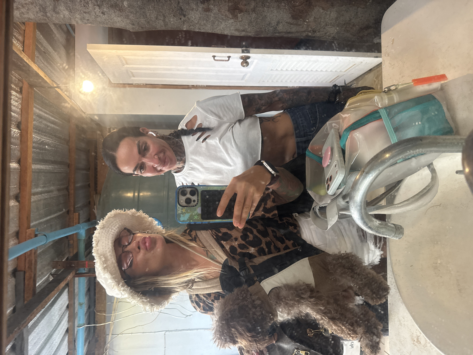 Two individuals are taking a mirror selfie in a bathroom, possibly in Choeng Thale, Thailand, judging by the presence of what appears to be a rough, unfinished ceiling and walls, and possibly a tree trunk visible in the background. The individual on the left is wearing a fluffy white bucket hat, sunglasses, and a leopard print top, and is holding a small brown dog in a carrier. The individual on the right is wearing a white crop top with ripped details and denim jeans. They are both holding a smartphone to take the picture. In the foreground, a sink and faucet are visible. The overall impression is a casual and somewhat rustic setting.