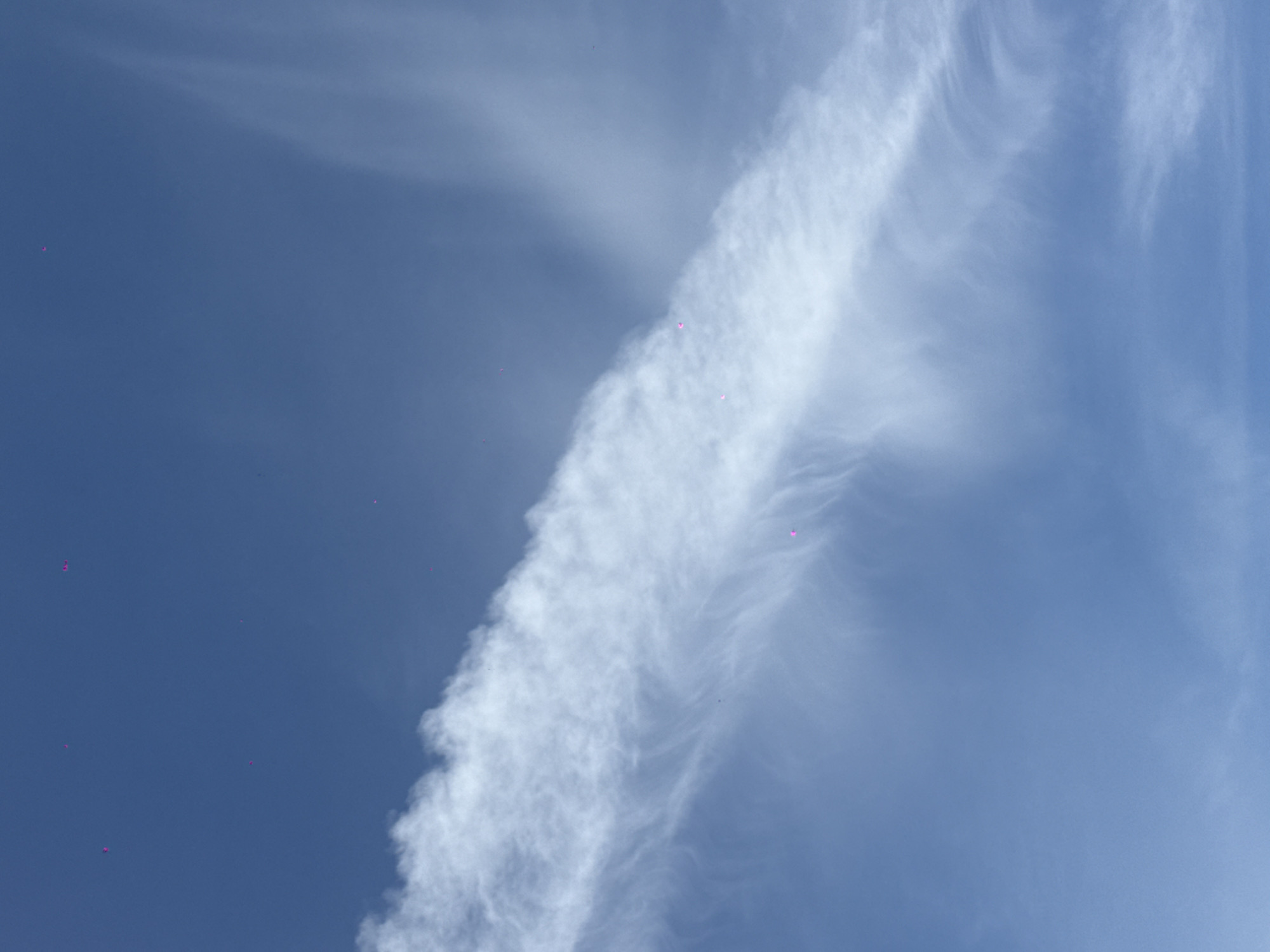 The image shows a daytime view of a blue sky with white clouds. A prominent linear cloud formation, possibly a contrail, stretches diagonally across the frame. There are also several small pink spots scattered throughout the image, possibly artifacts from the camera or sensor. The location of Khimki, Russia, is not evident in the image.