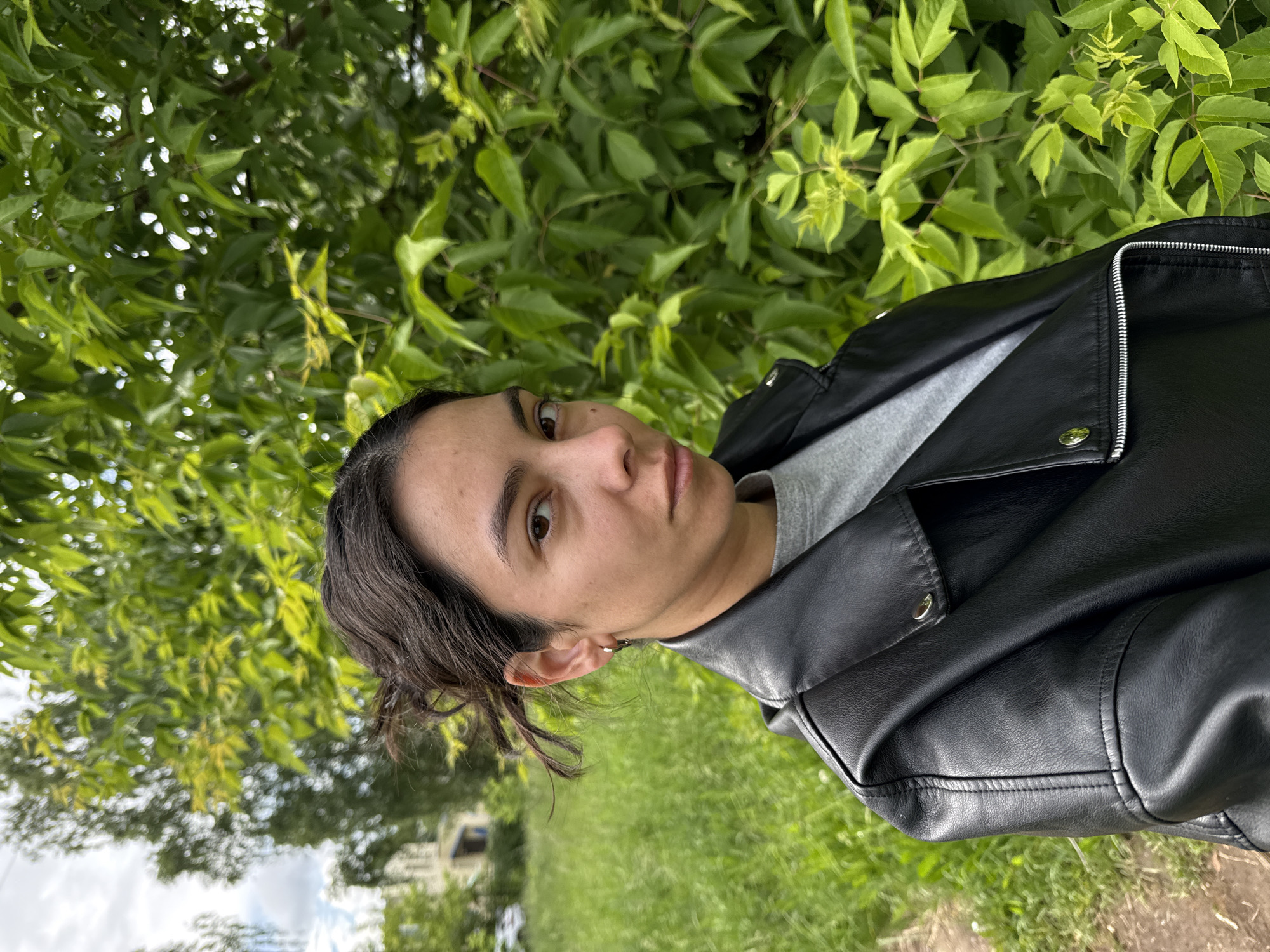 A waist-up portrait shot in Chaikovsky, Russia, features a person looking directly at the camera with a neutral, calm expression. The subject, appearing to be female, has dark hair pulled back, dark eyes, and full lips, with a small hoop earring visible in her left ear. She is wearing a black faux leather jacket with prominent silver zippers and snap buttons, over a grey t-shirt.
The setting is outdoors, dominated by lush, vibrant green foliage that fills the background, suggesting a natural environment such as a park or garden. The leaves are well-lit, indicating a daytime scene, possibly under an overcast sky or in a shaded area given the soft, even lighting and absence of harsh shadows. Beyond the immediate greenery, blurry hints of distant structures and a cloudy sky can be seen in the lower left portion of the background. The person is posing for the photograph. There is no visible text in the image.