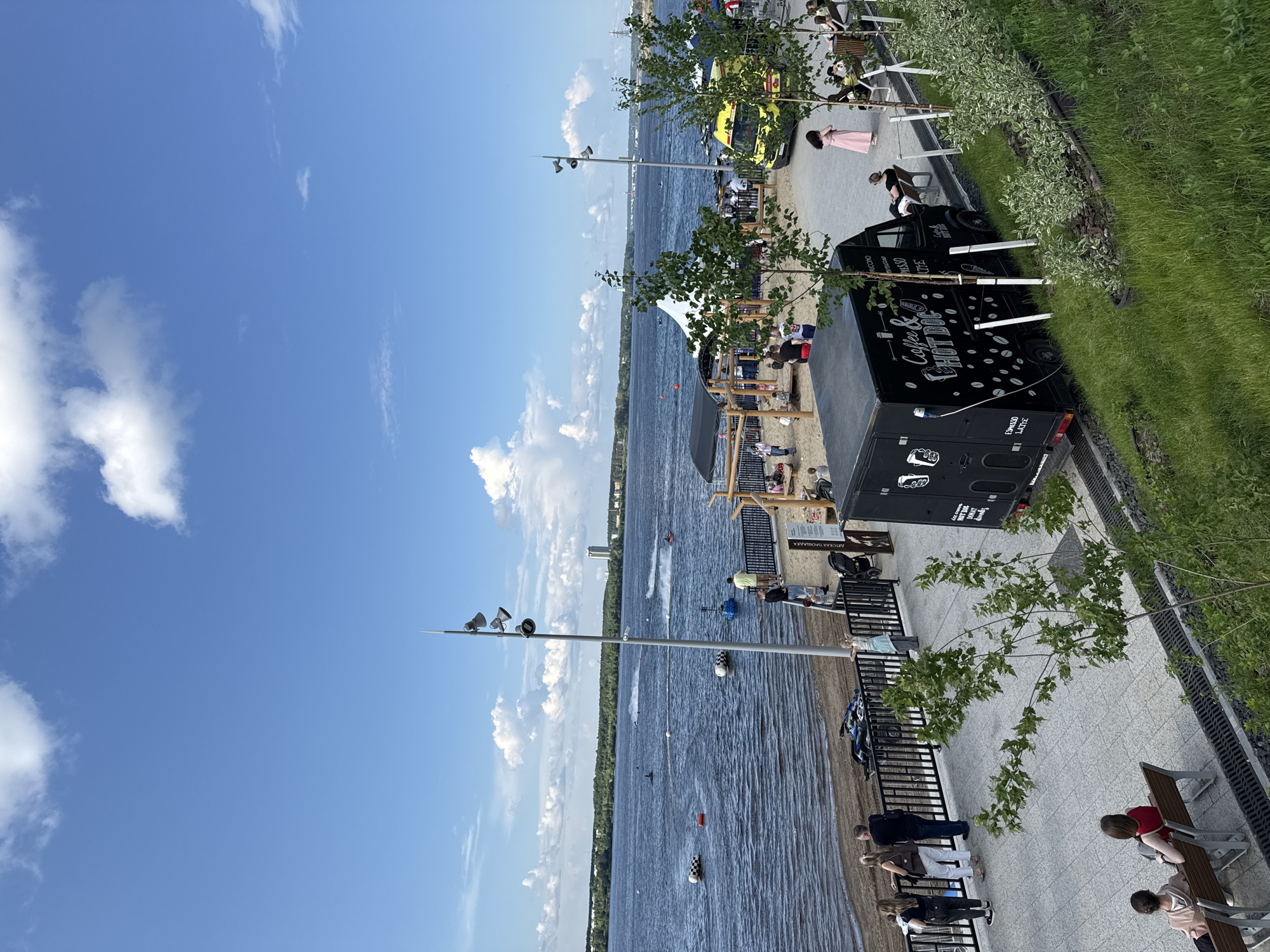 This is an exterior, eye-level, full shot of a waterfront area in Perm, Russia, on a bright, cloudy day. In the foreground, a grassy embankment slopes down to a paved walkway. A black food truck with white lettering that reads "Coffee & HOT DOG" is parked on the walkway. Several trees with green leaves are scattered throughout the scene. A group of people are seated on benches on the right side of the frame, facing away from the camera. Further back, the walkway borders a sandy beach area where several children and adults are present. Beyond the beach is a wide, dark blue river with multiple jet skis and small boats visible. The far bank of the river is lined with trees, and a tall, modern building is visible in the distance. The sky is a bright blue with scattered white clouds.